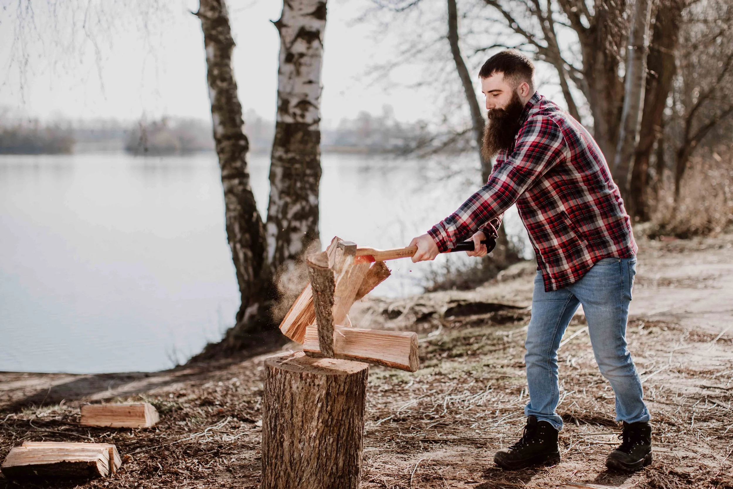 A man with a beard in a plaid shirt and jeans chopping wood with an axe by a lake, surrounded by trees.