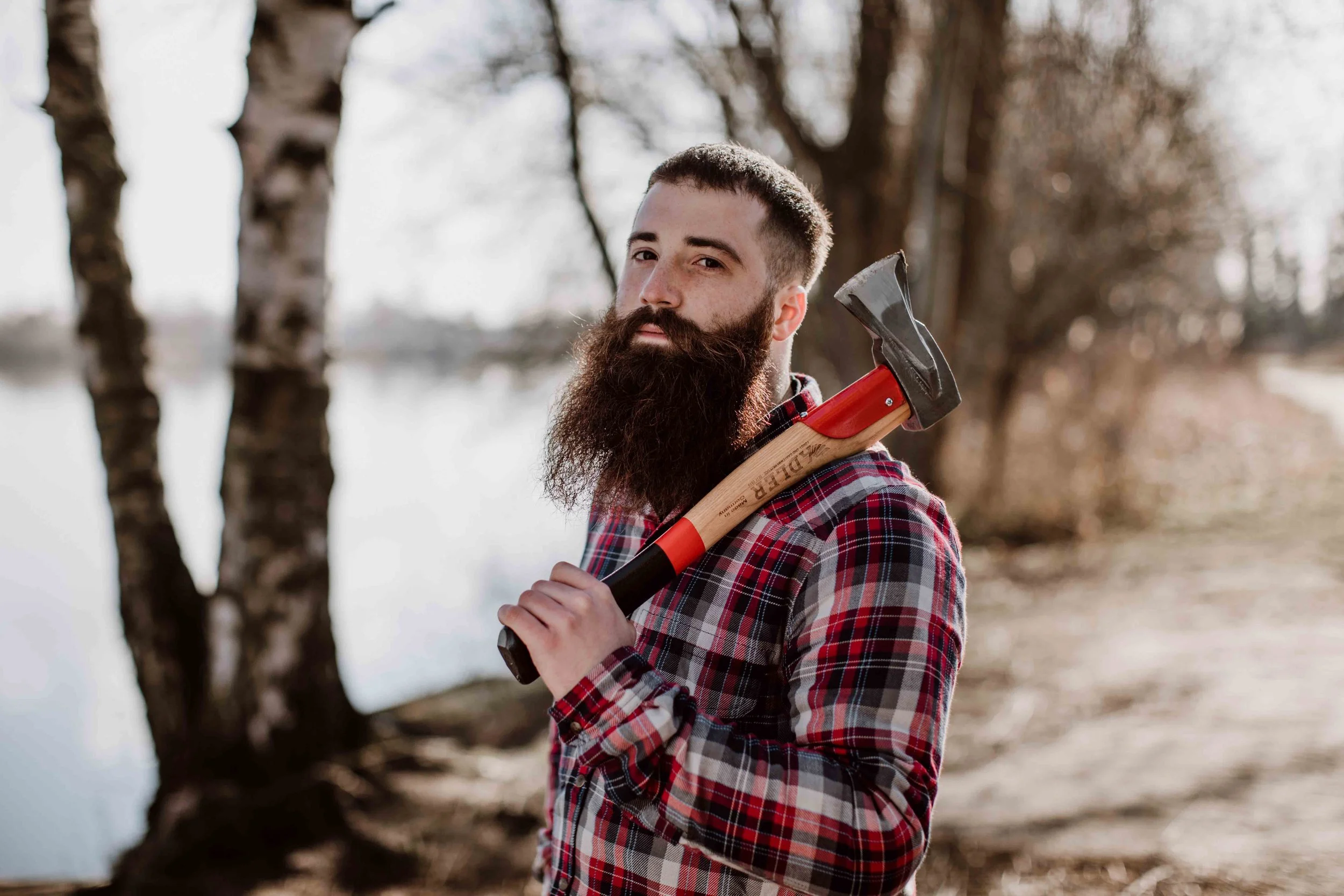 A man with a beard and short hair, wearing a plaid shirt, holding a hatchet over his shoulder, standing outdoors near a body of water with trees in the background.