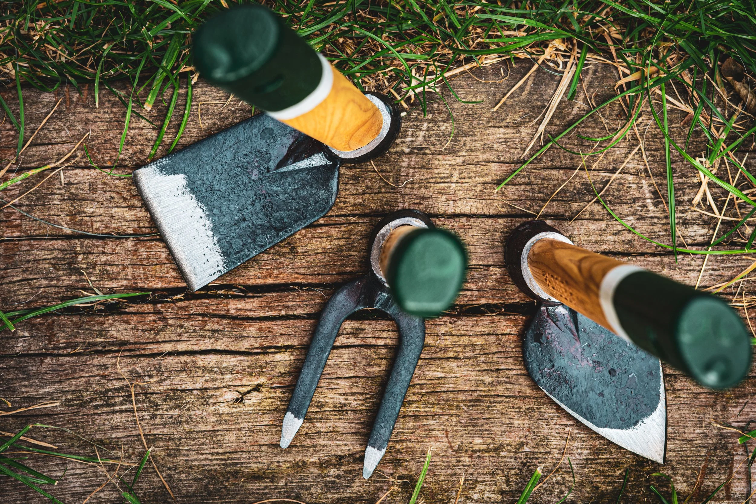 A set of gardening tools, including a small hand saw, a pruner, and a trowel, resting on a weathered wooden surface with grass around.