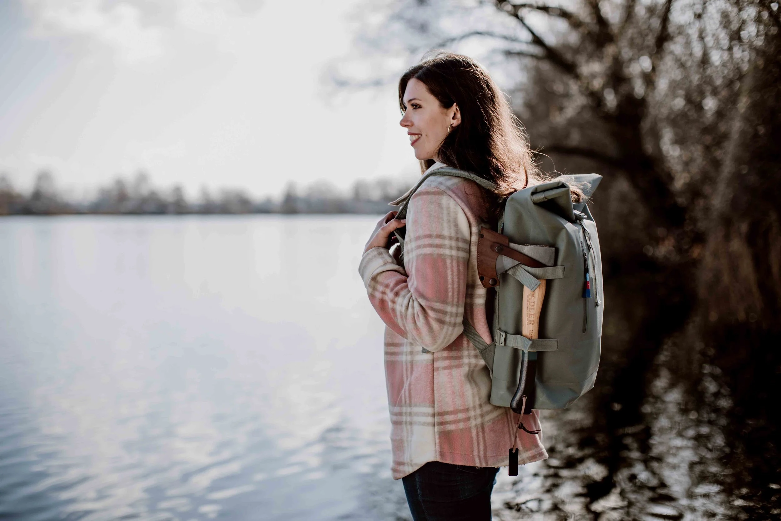 A woman with dark brown hair, wearing a plaid coat, standing by a lake with trees in the background, carrying a large green backpack.