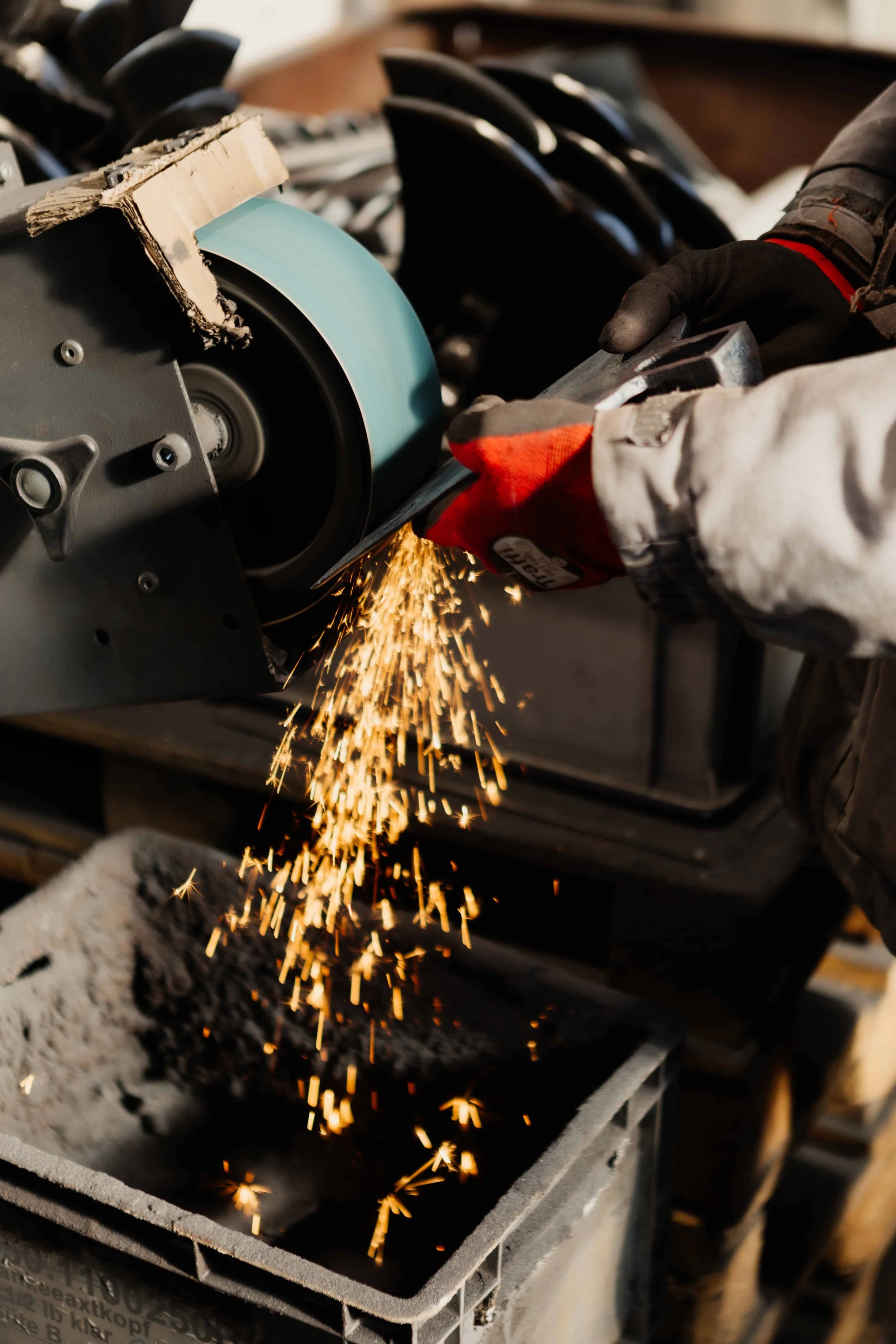 A person wearing protective gloves uses a grinding tool to sharpen a metal blade, with sparks flying from contact with a grinding wheel.