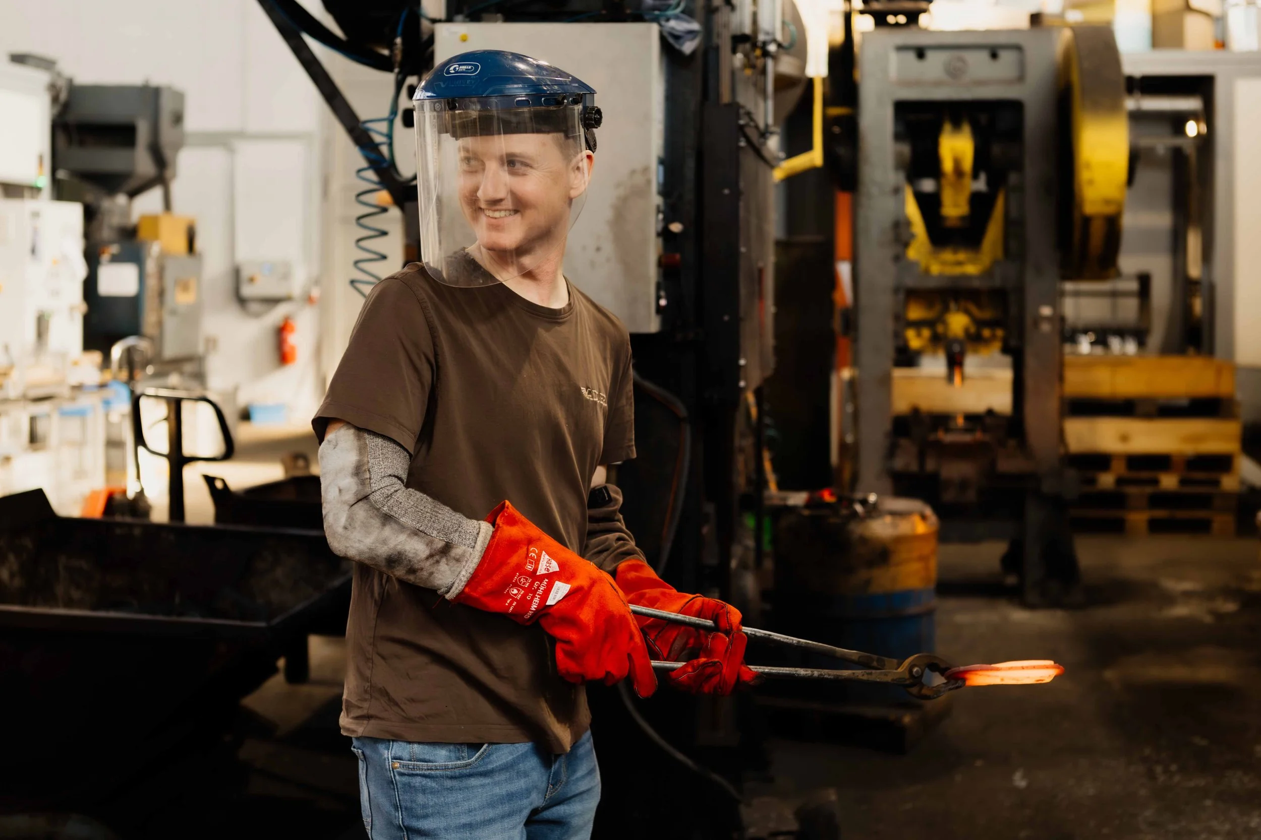 A young man wearing a face shield, brown t-shirt, jeans, and red gloves standing in a workshop holding metal tongs with a glowing hot piece of metal.