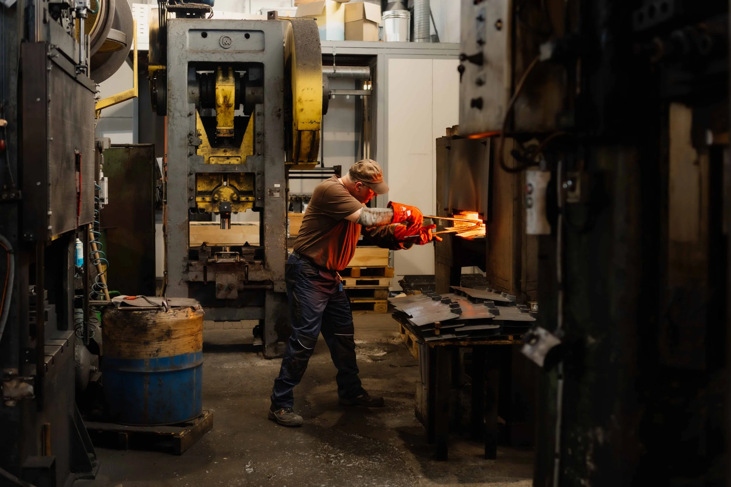 A worker in a manufacturing plant is working with heated metal, using tongs and safety gear, with industrial machinery around him.