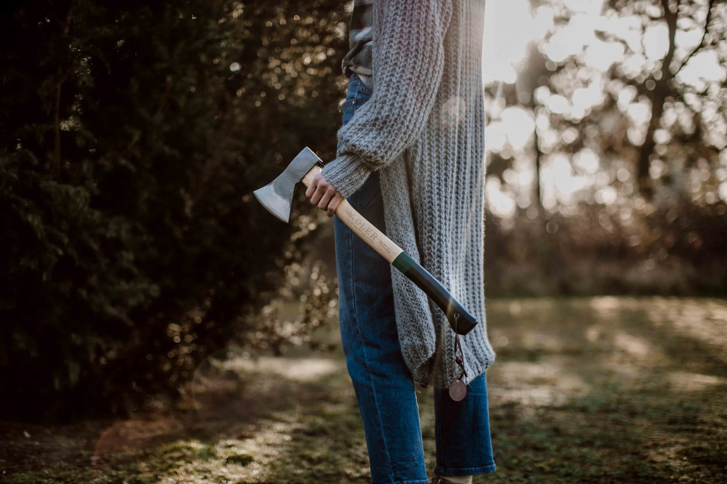 Person standing outdoors on a sunny day, holding an axe in their right hand, wearing a long knit cardigan and jeans.