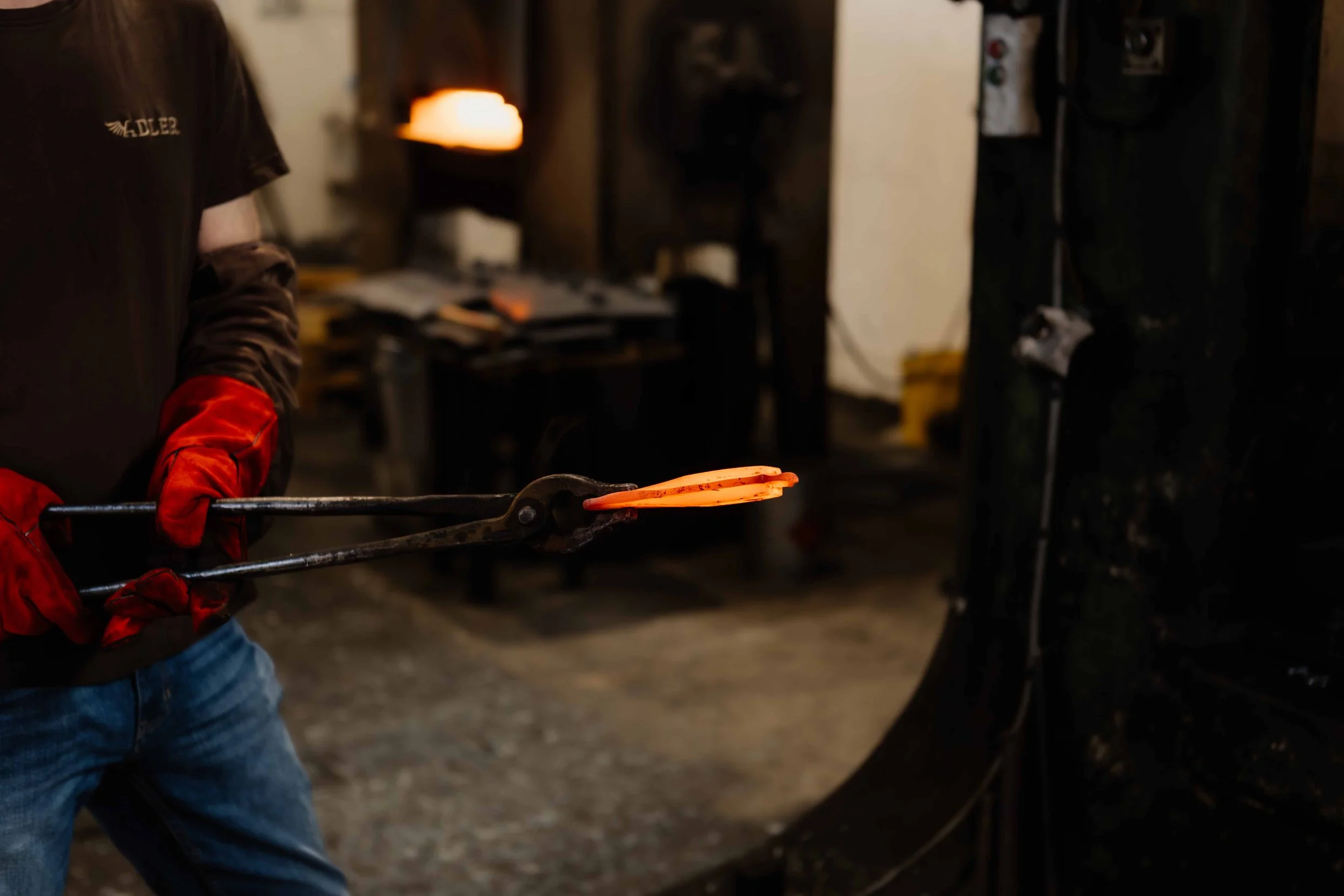 A person wearing red heat-resistant gloves holding black tongs with glowing hot metal being shaped in a blacksmith forge.