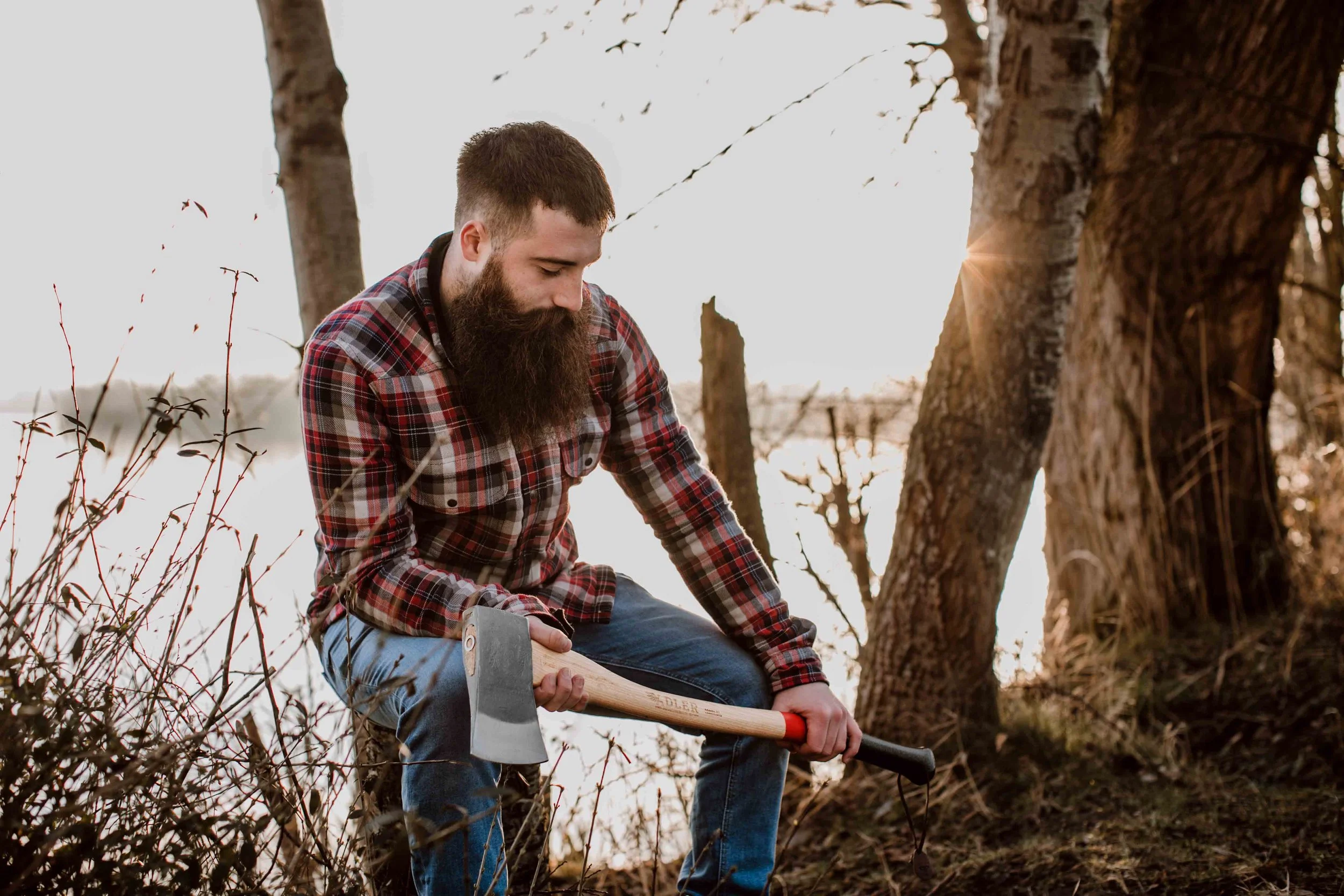 A man with a beard, wearing a red plaid shirt, sitting outdoors by trees, holding an axe.