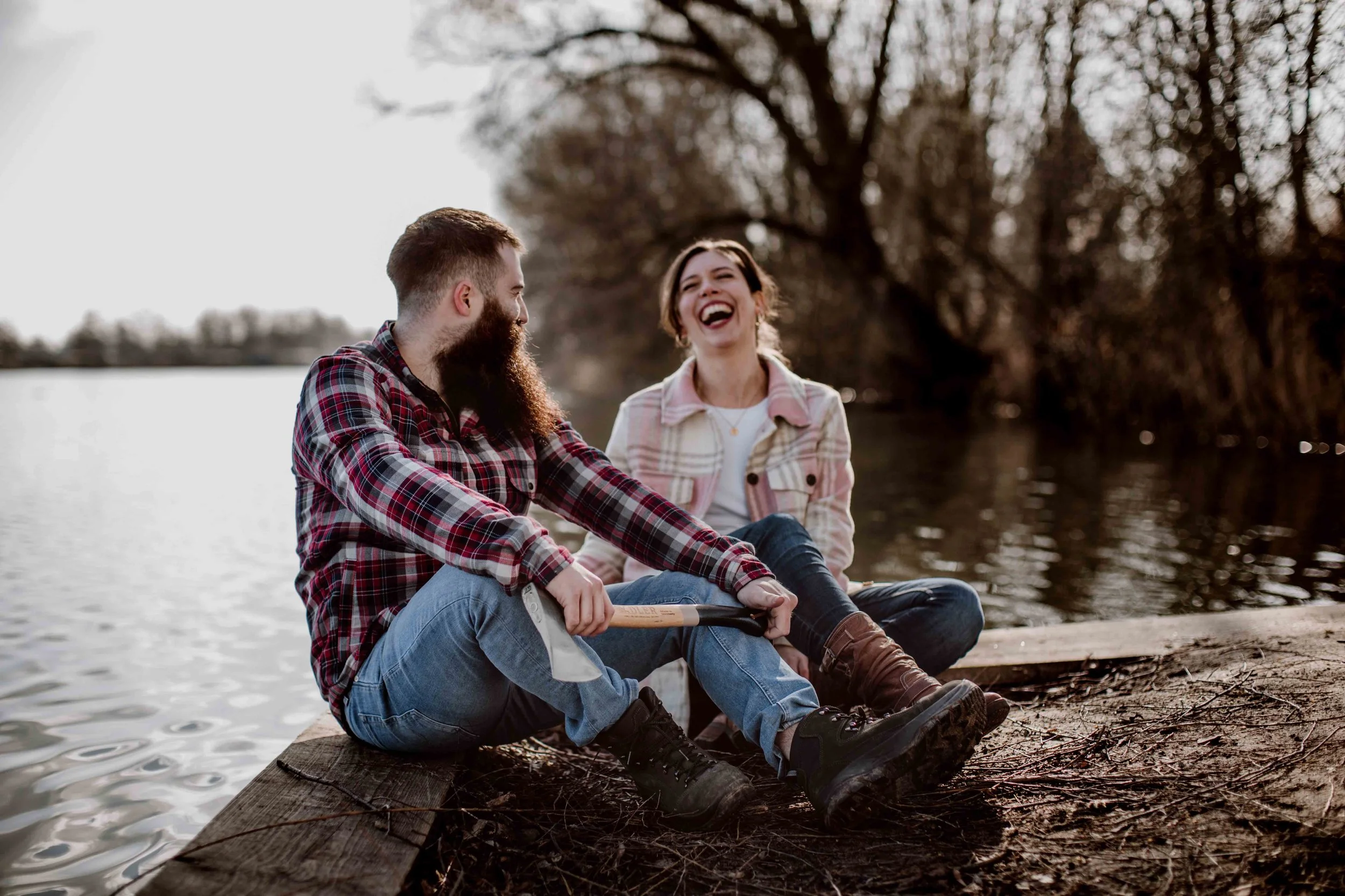 A man and a woman sitting on a dock by a lake, enjoying each other's company and laughing, with trees in the background.