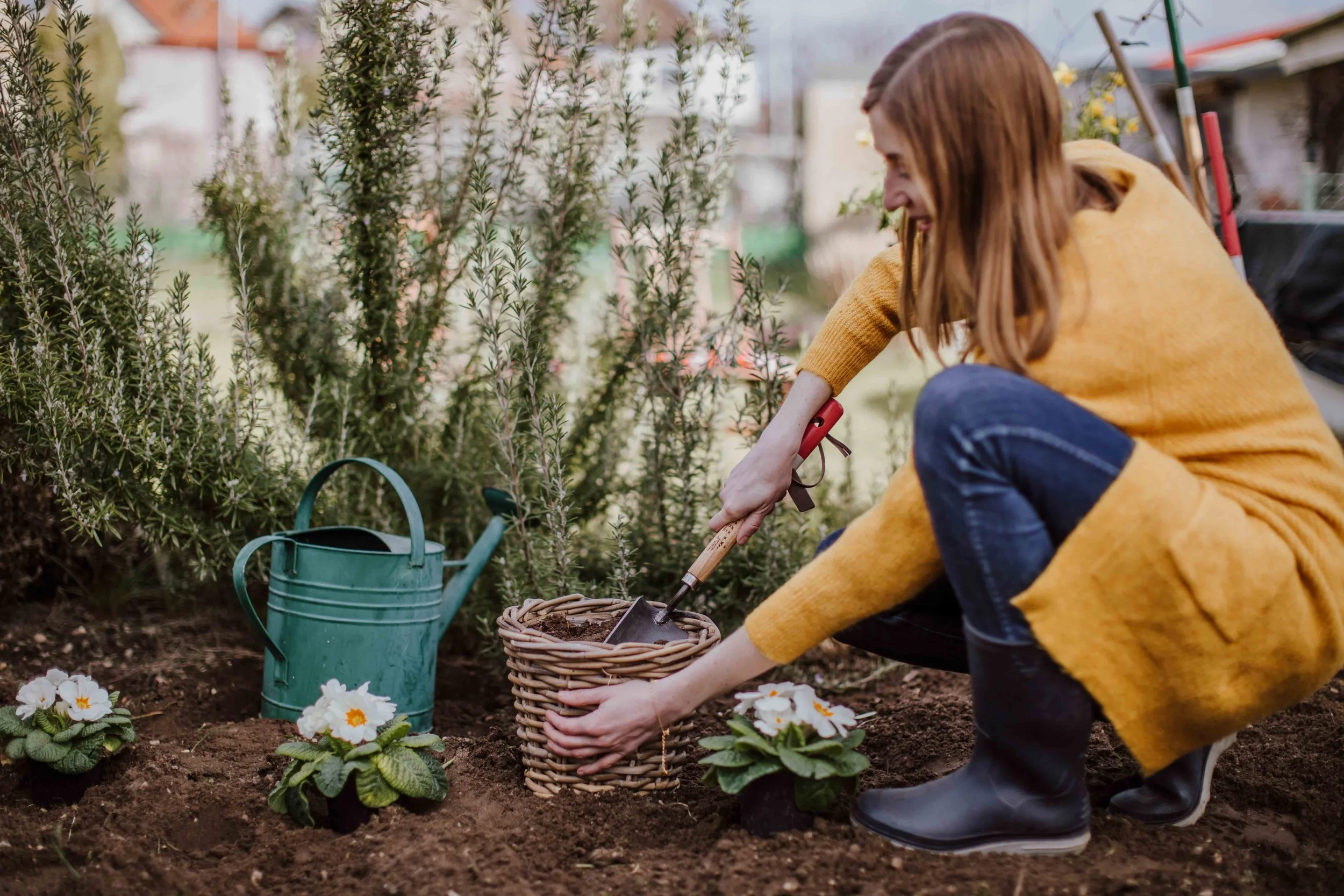 A woman planting flowers in a garden, wearing a yellow coat, black boots, and using a garden trowel, with a green watering can nearby.