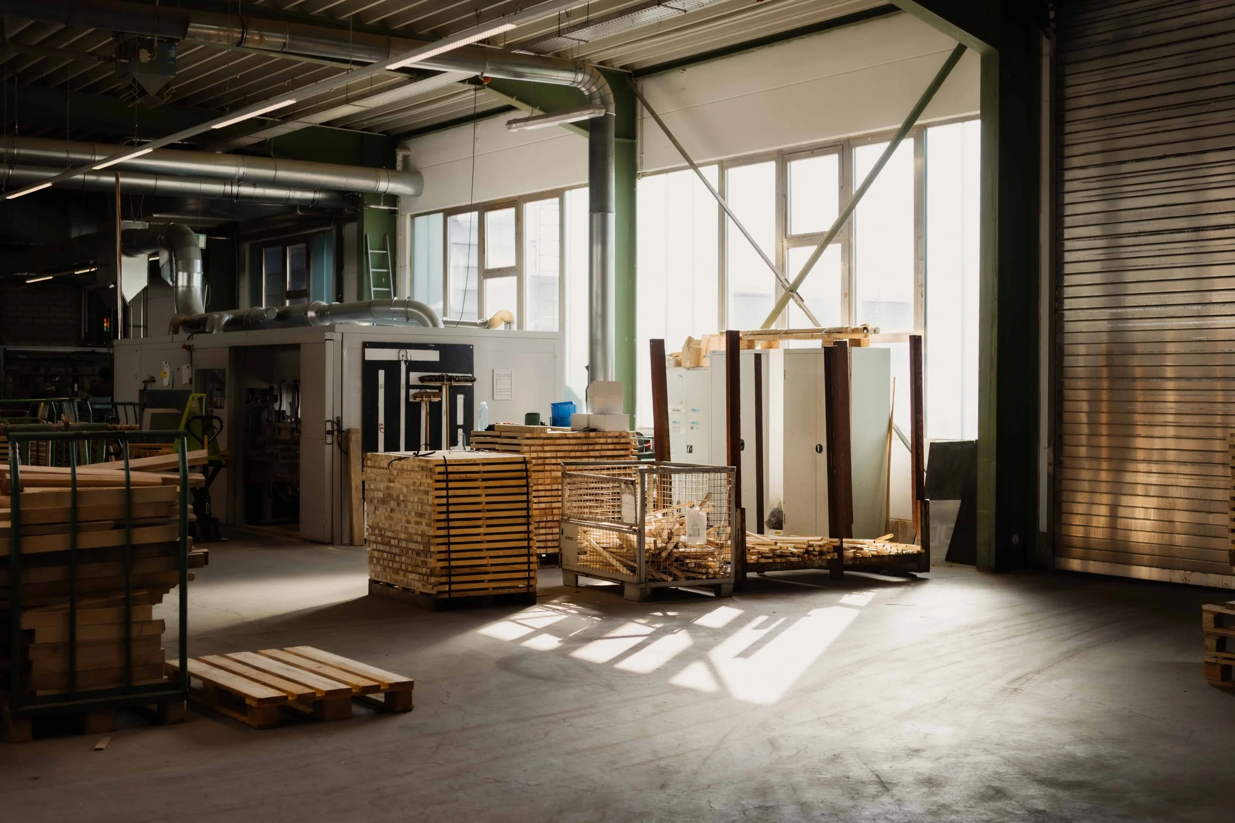 Interior view of a woodworking workshop with stacks of wood and machinery near large windows