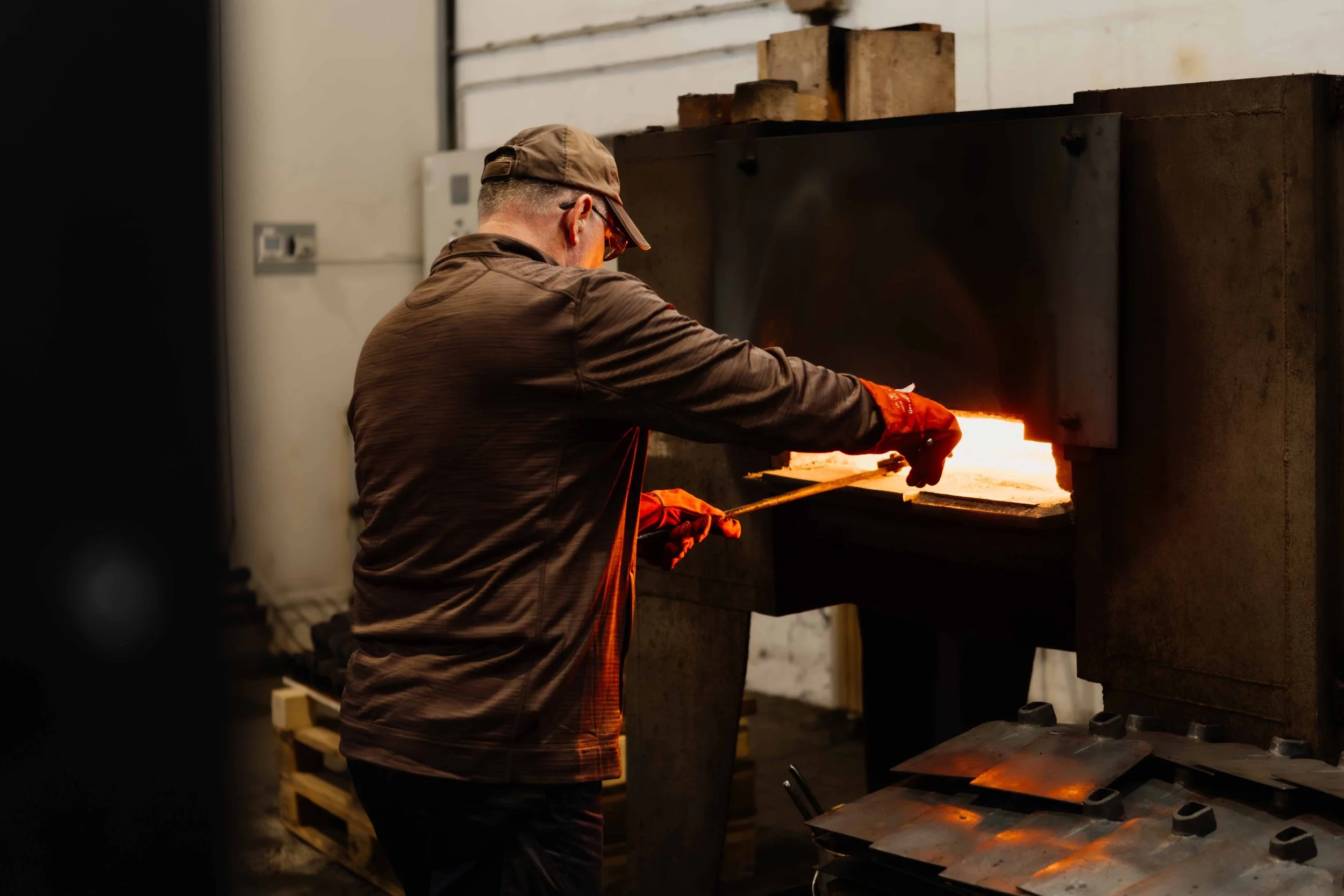 A worker in safety gloves and glasses is heating and manipulating metal in a large industrial furnace.