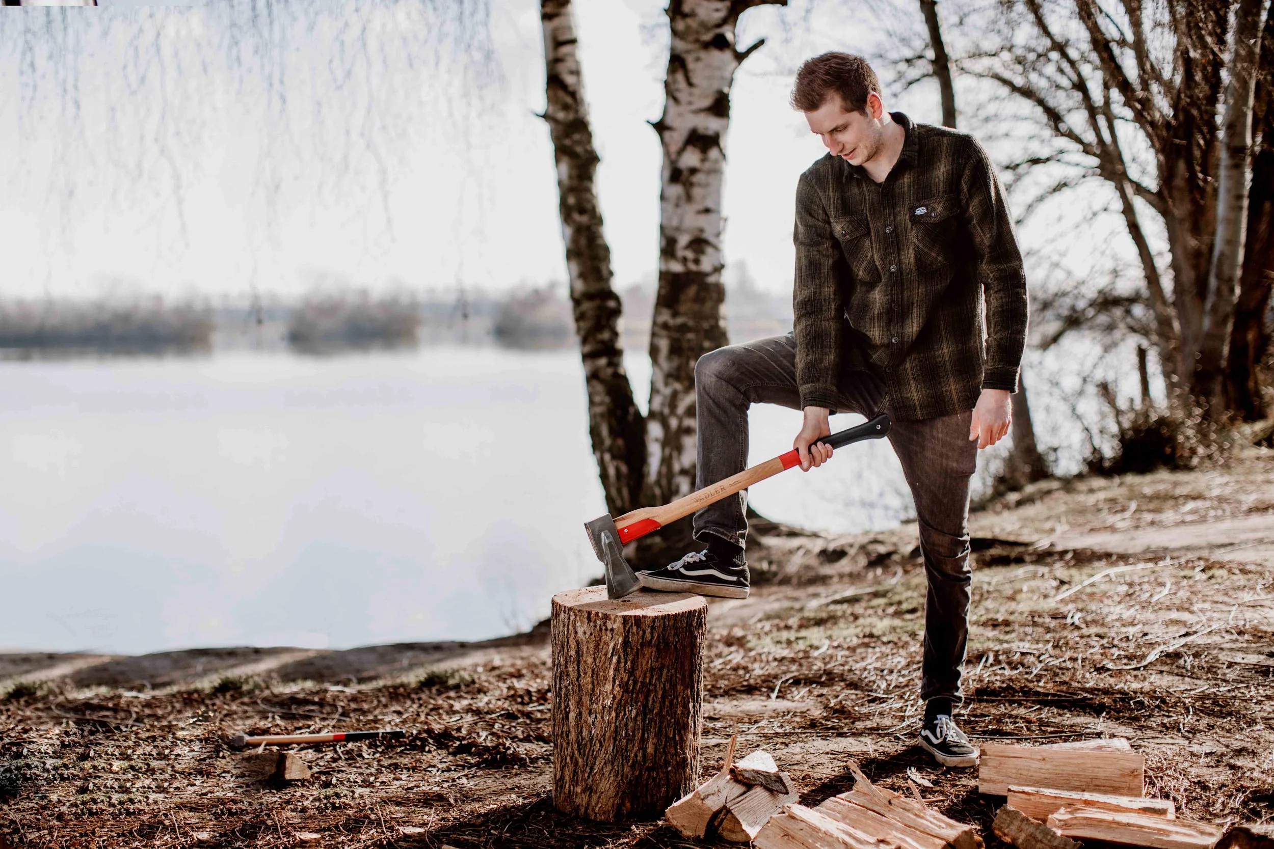 A young man chopping wood with an axe outdoors near a lake or river, with trees in the background.