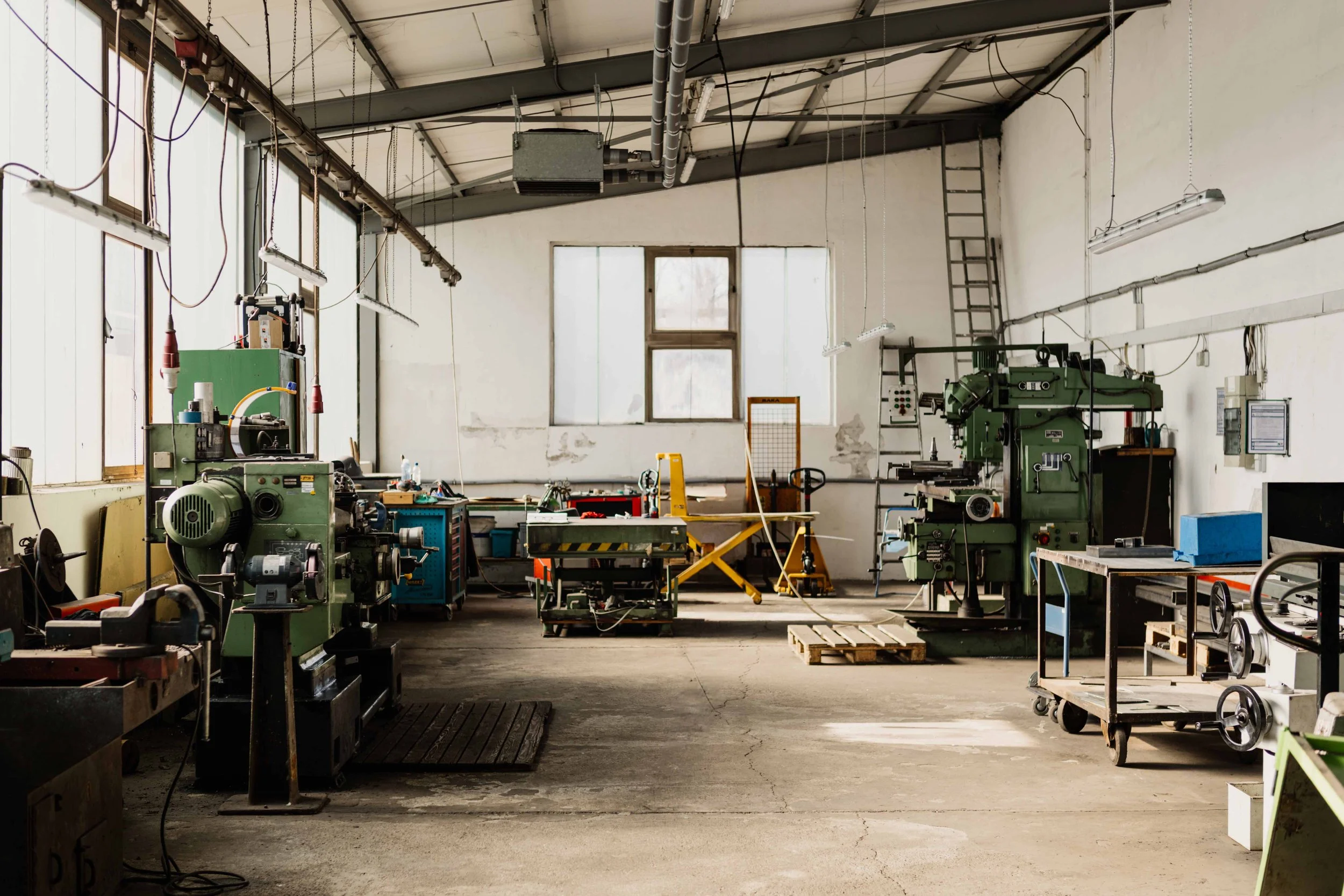 An empty industrial workshop with various green machines, worktables, and tools, illuminated by hanging lights and natural light from large windows.