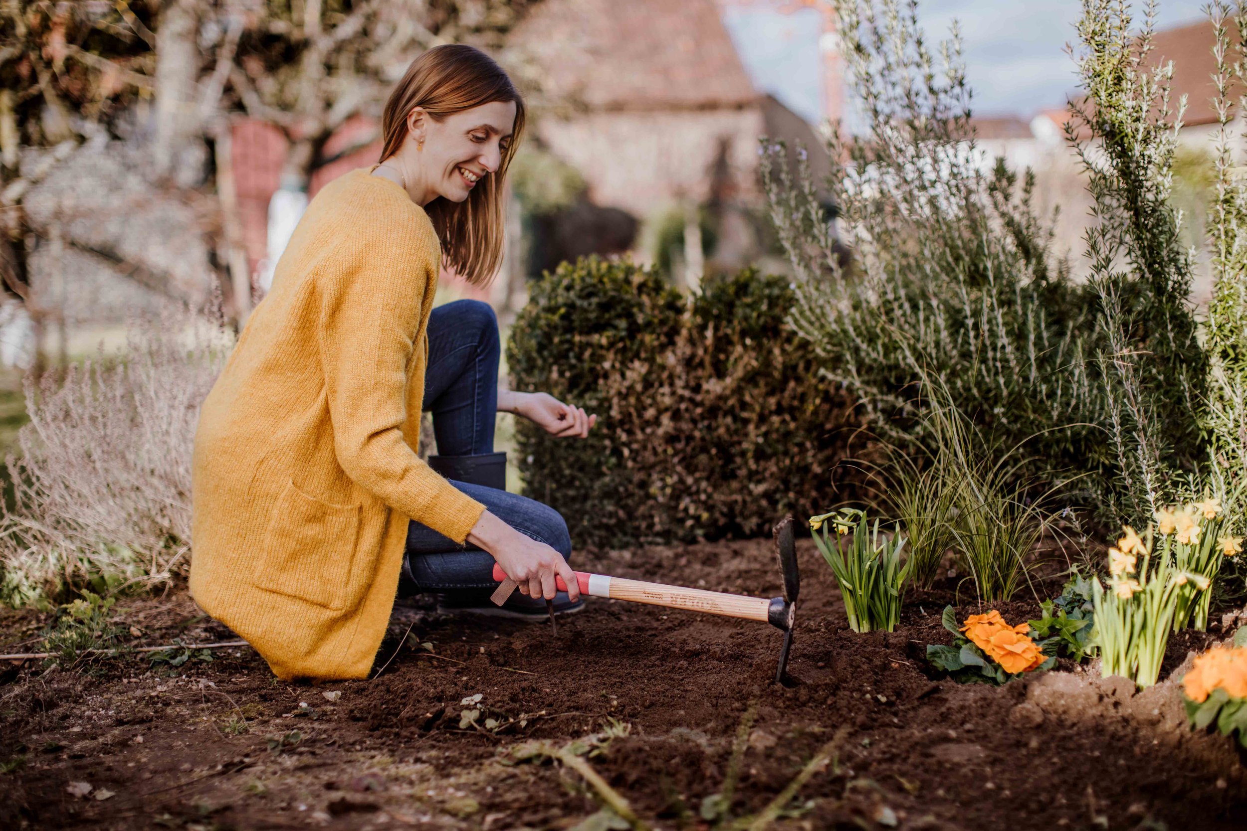 A woman in a yellow sweater and jeans kneels in a garden, planting flowers with a small gardening shovel, surrounded by shrubs and blooming flowers.