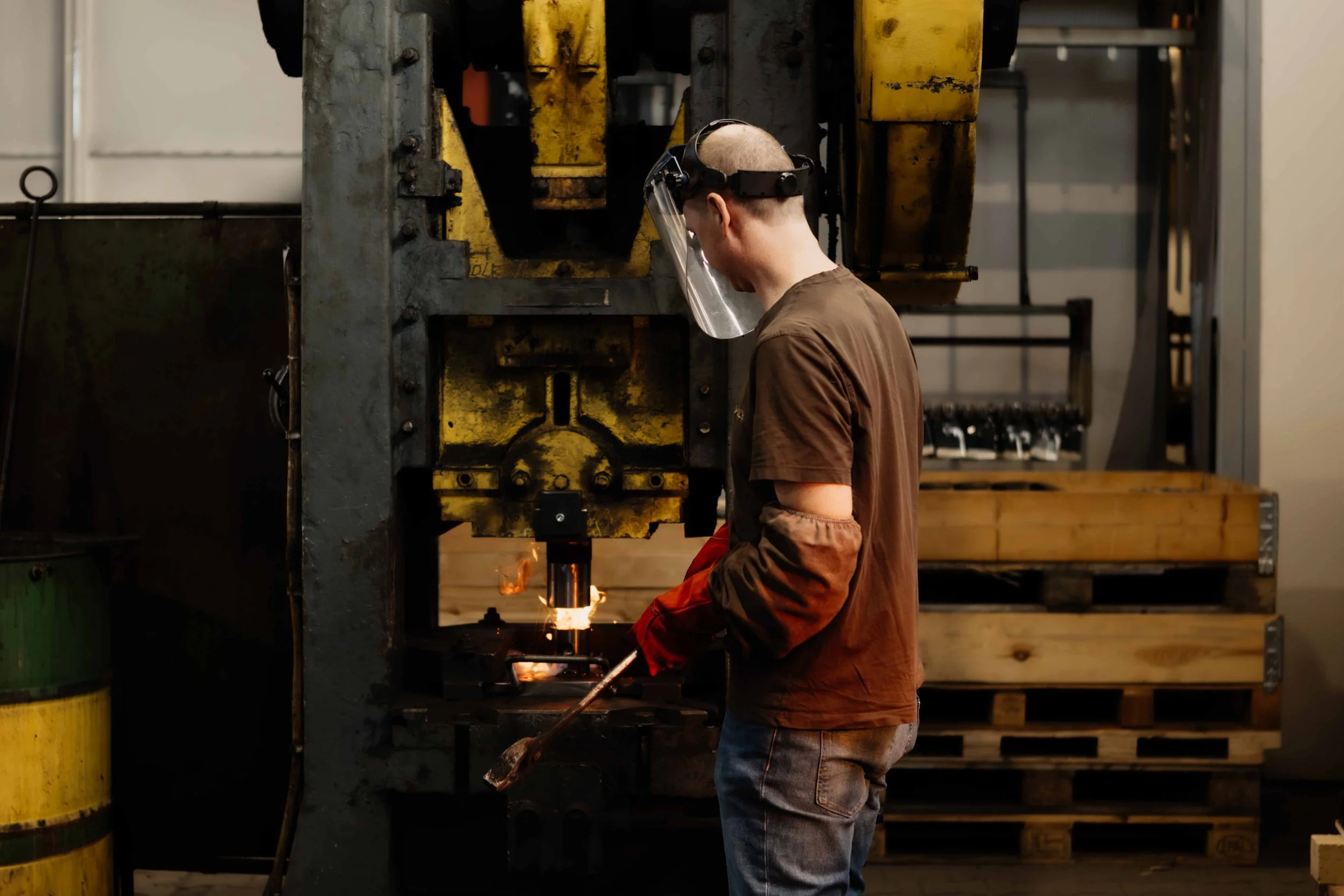 A worker wearing safety goggles, a face shield, and gloves operating large industrial machinery with sparks flying, in a factory setting.
