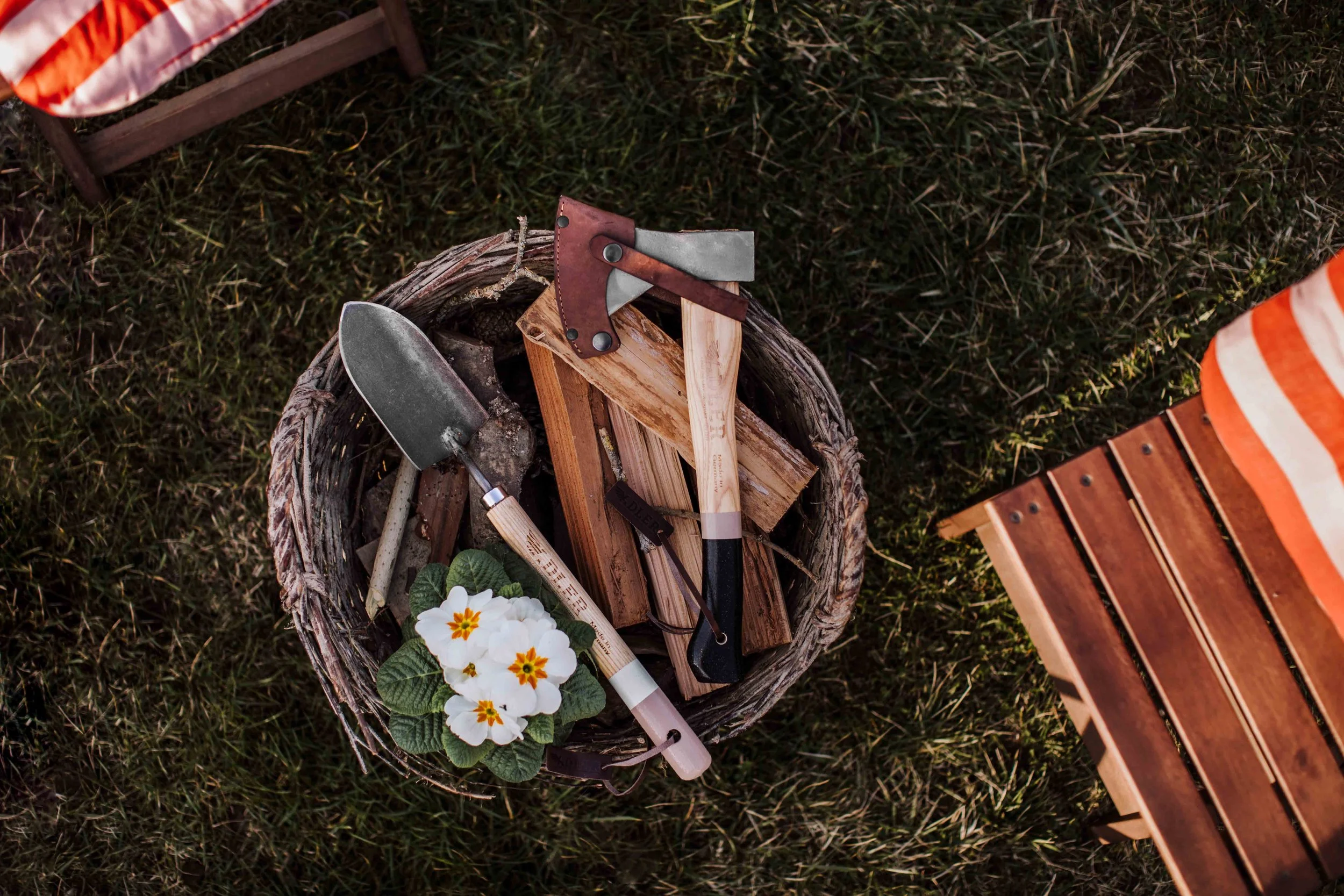 A basket filled with gardening tools, including a small shovel, a hoe, and a pair of pruning shears, along with a white flowering plant with green leaves, is placed on grass next to a wooden bench and some outdoor furniture.