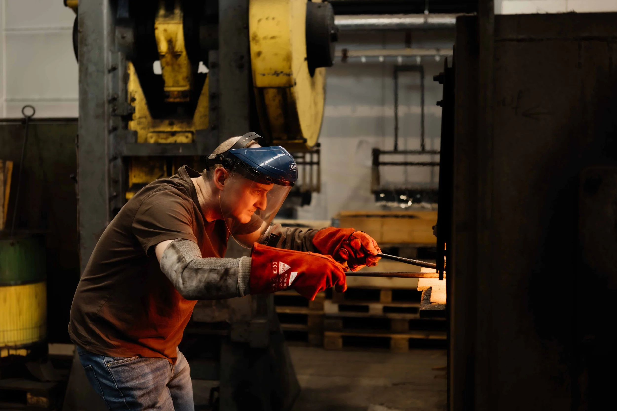 A man wearing a welding helmet, safety gloves, and protective arm sleeves welding a metal piece indoors in an industrial workshop.