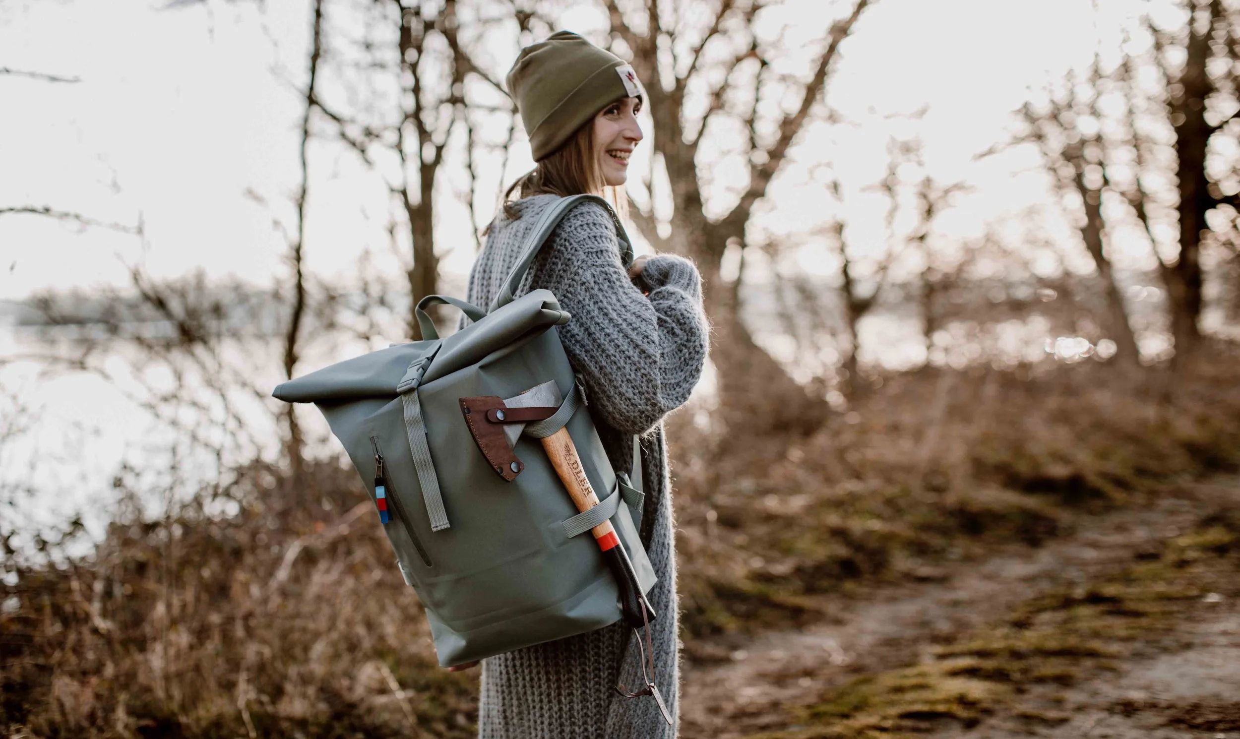 Woman in a gray knit sweater and beanie hat walking outdoors with a gray backpack on a forest trail during daytime.