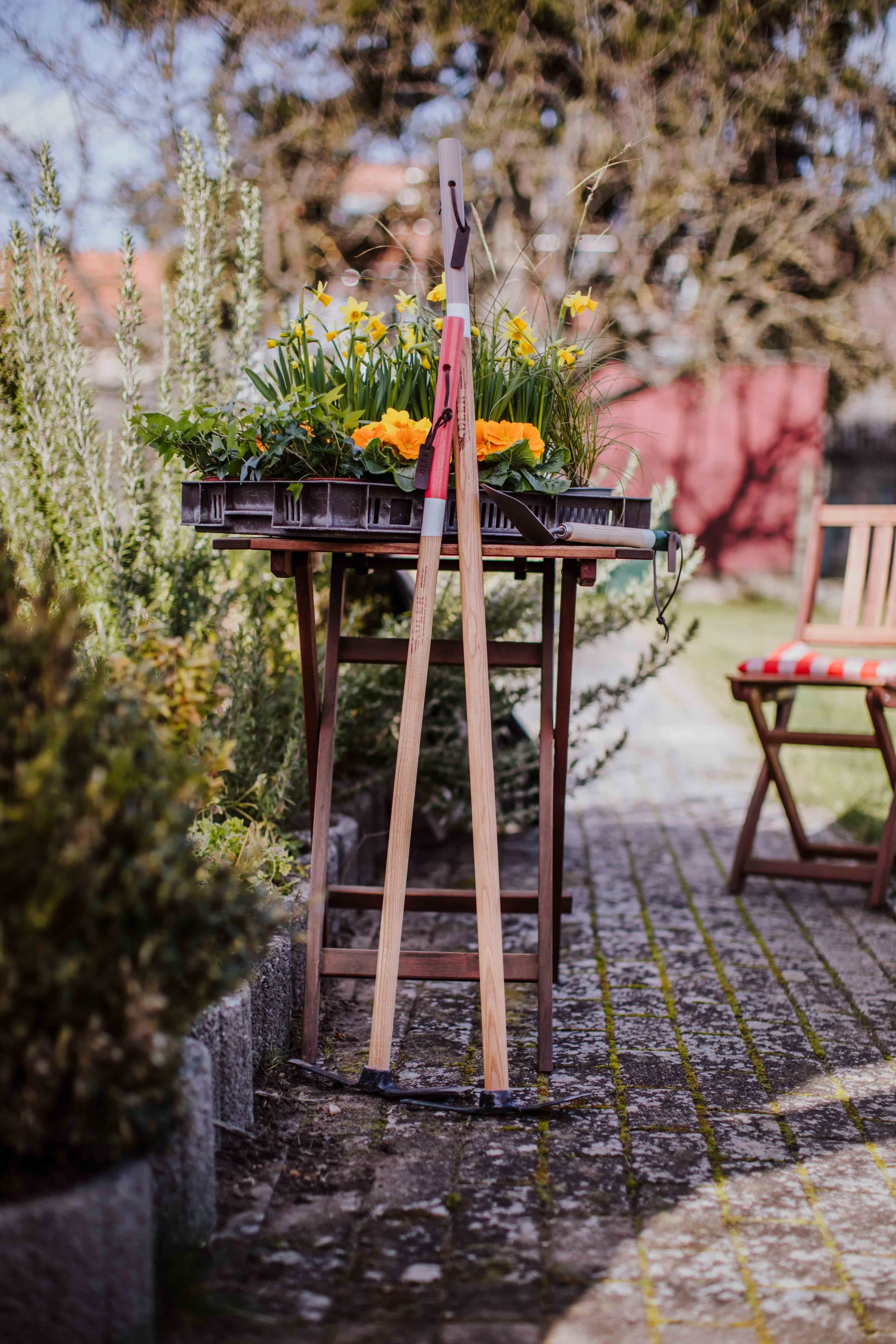 Garden tools leaning against a small wooden table with potted flowers and plants, with a pathway and outdoor furniture in the background.