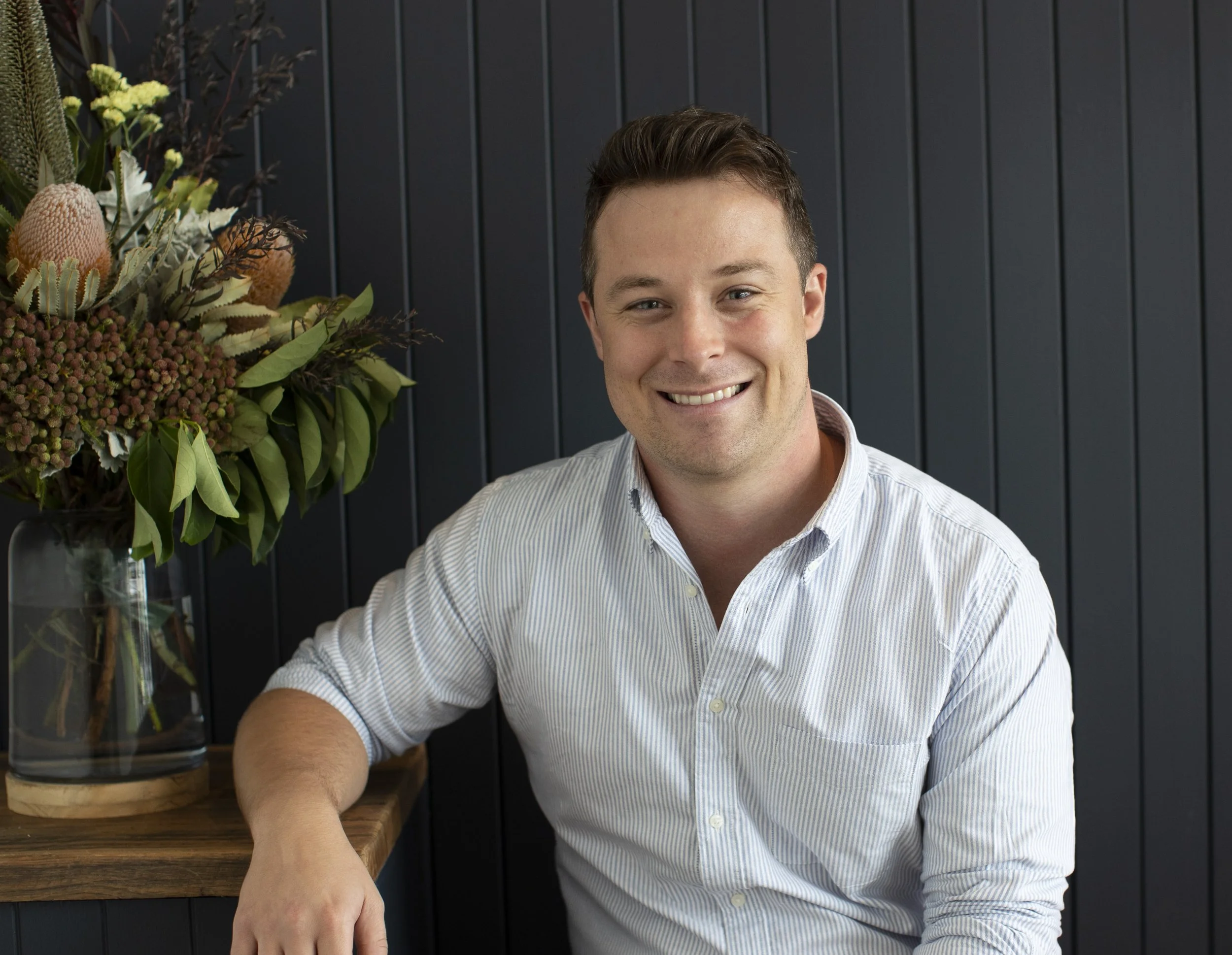 A smiling man sitting at a table, wearing a light blue striped button-up shirt, with a large bouquet of flowers and green foliage in a glass vase on the table behind him, against a dark paneled wall background.