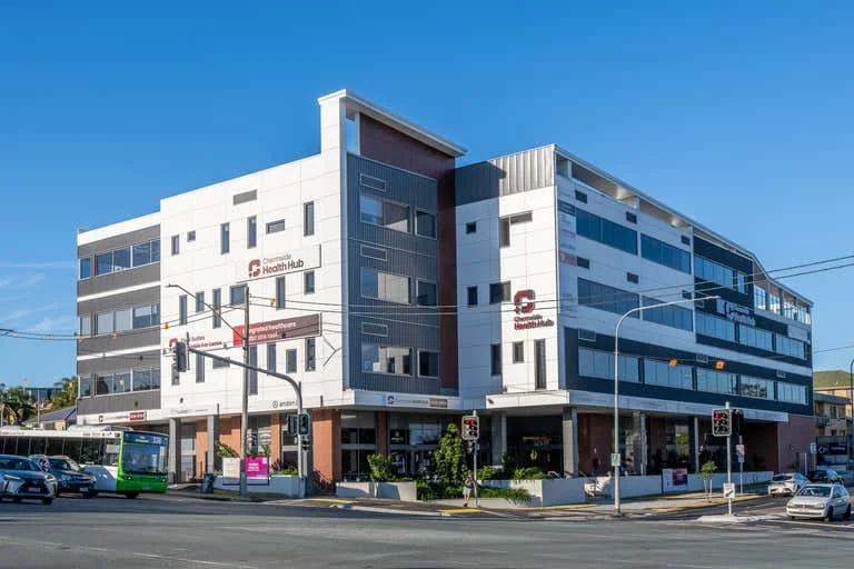 Modern multi-story healthcare building with signage for Chermside Health Hub, situated on a busy street with cars and a green bus, under a clear blue sky.