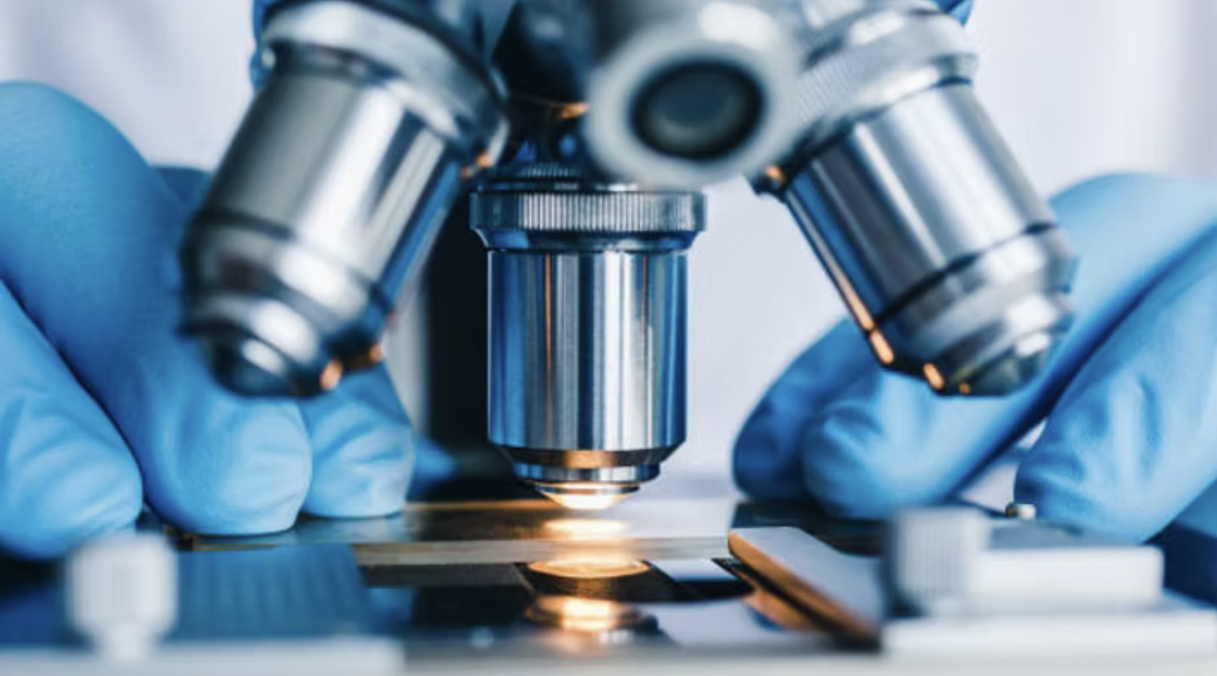 Close-up of a microscope with a scientist or technician wearing blue gloves examining a slide in a laboratory.