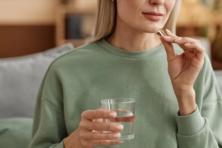 A woman holding a glass of water and a pill near her mouth, preparing to swallow.
