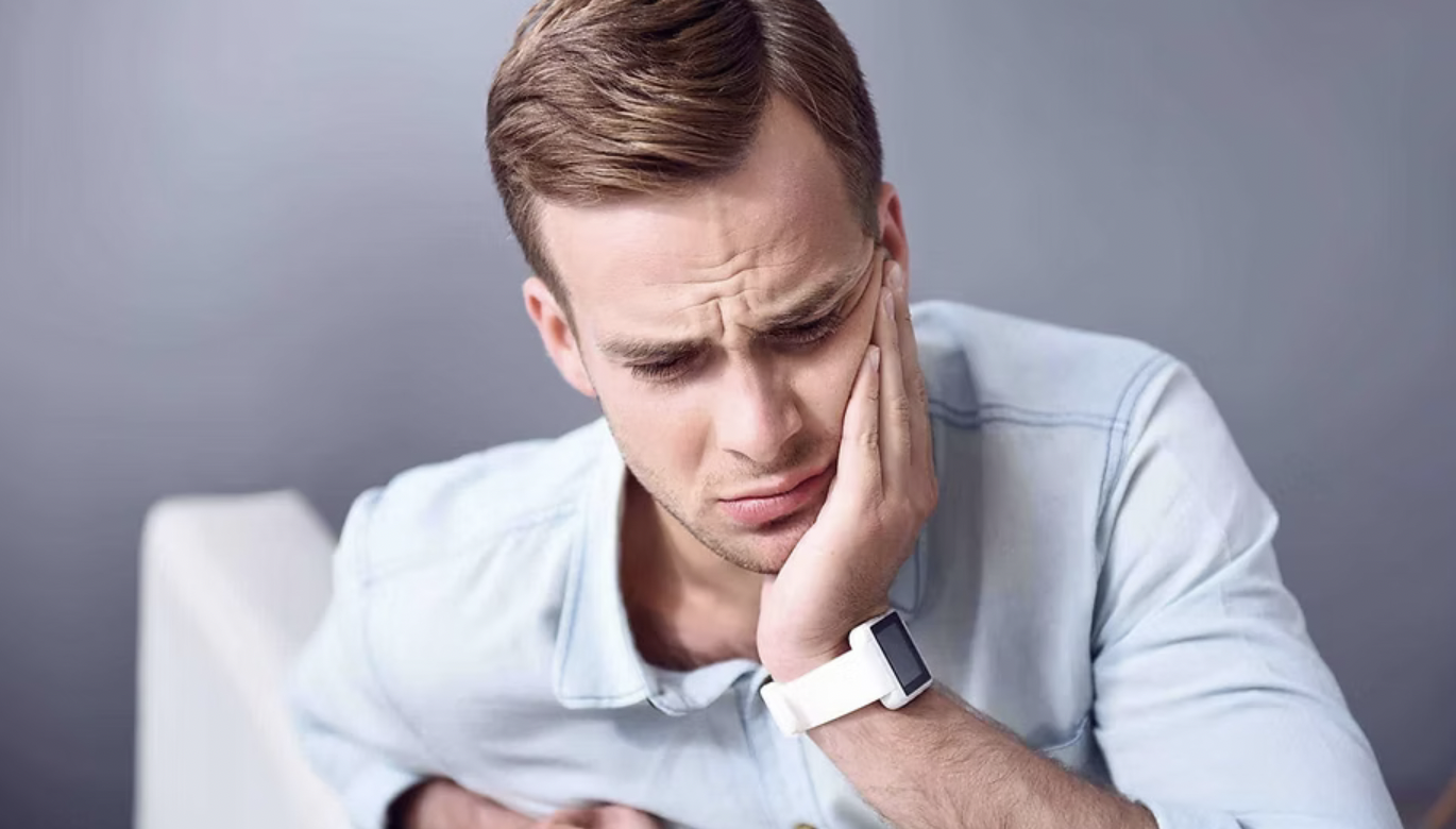 Man with brown hair holding his jaw in pain, wearing a white shirt and smartwatch, in a gray background.