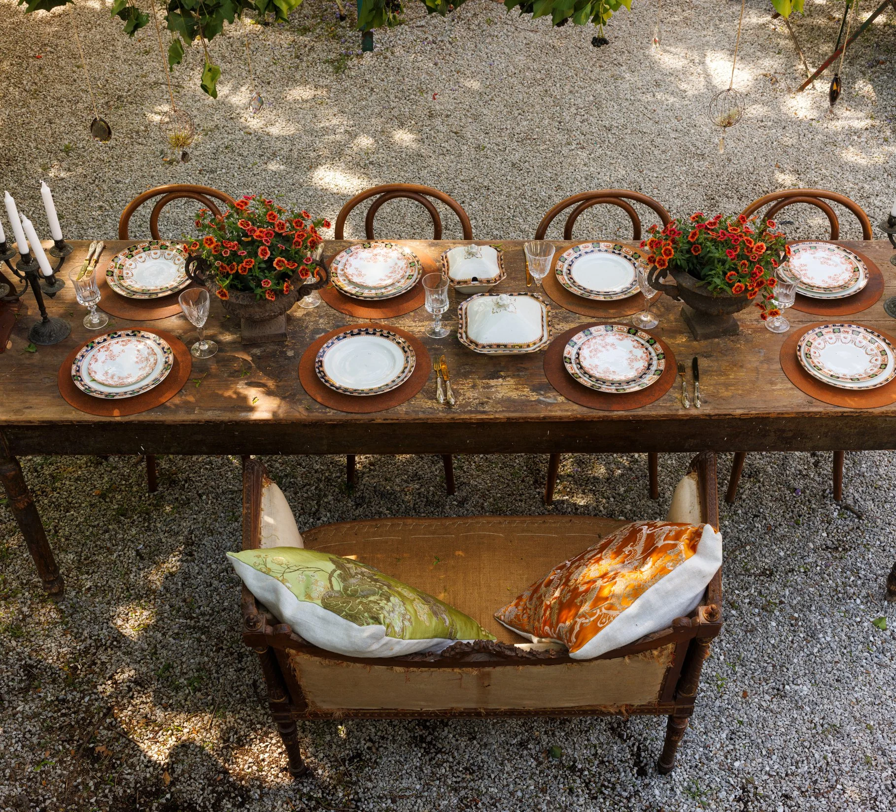 A rustic outdoor dining table set with floral-patterned plates, wine glasses, and gold flatware, decorated with potted red flowers and candles, viewed from above on a gravel surface with a vintage sofa with decorative pillows at one end.