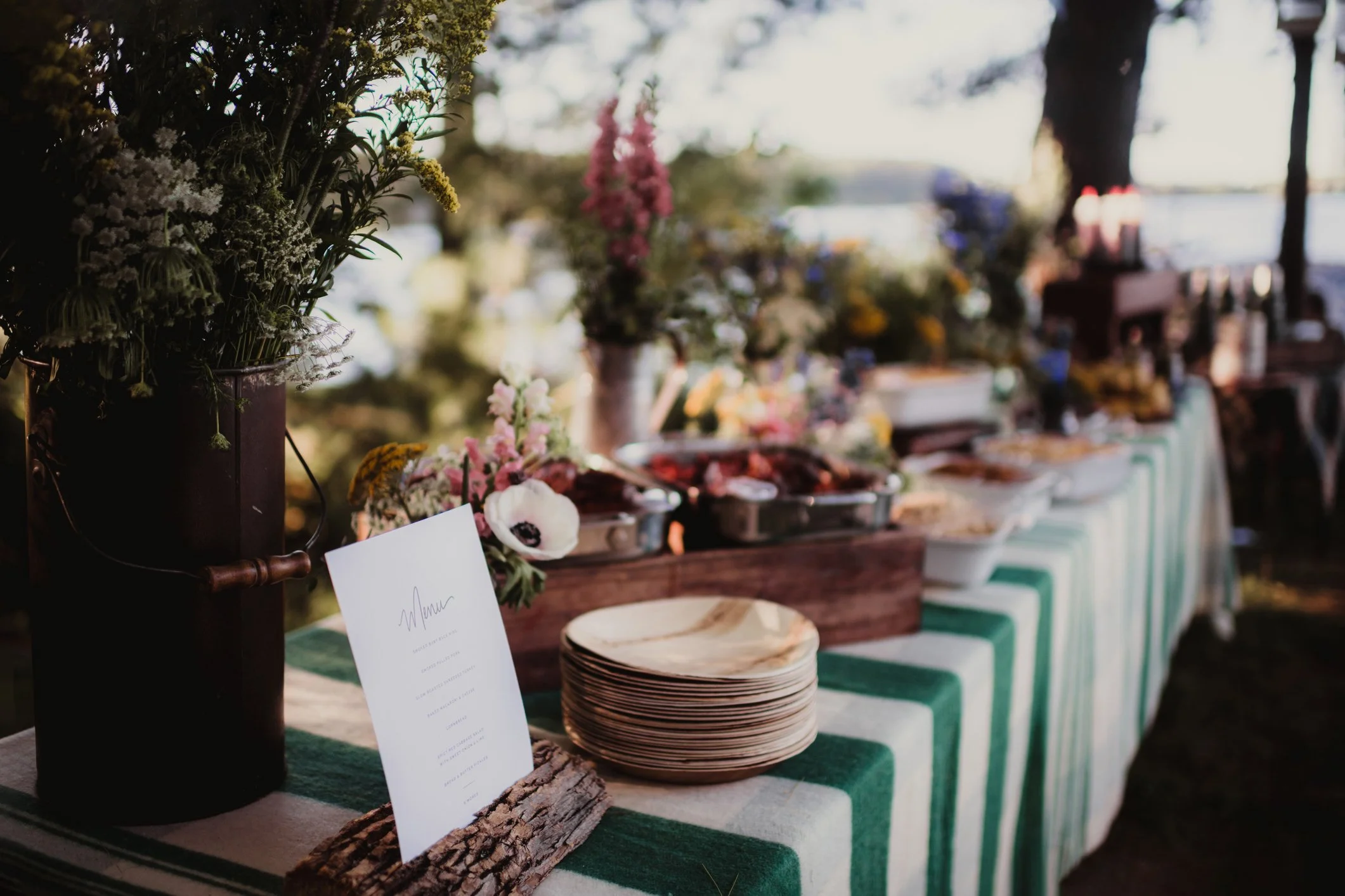 A buffet table outdoors decorated with green and white striped tablecloths, floral arrangements, plates, and various food dishes, with trees and an open sky in the background.