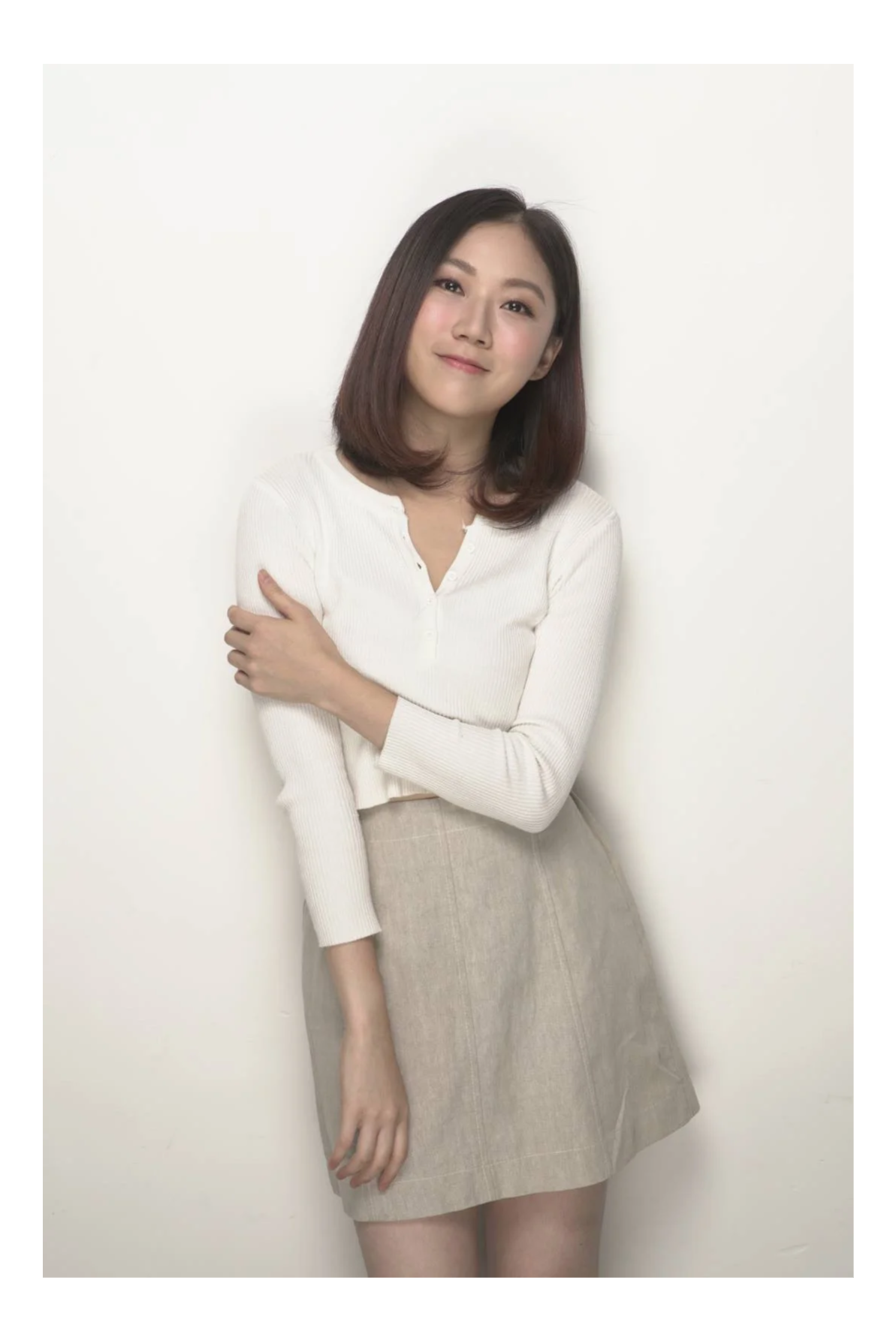 A woman with shoulder-length dark hair, wearing a white long-sleeve top and a beige skirt, standing against a plain white wall, smiling softly.
