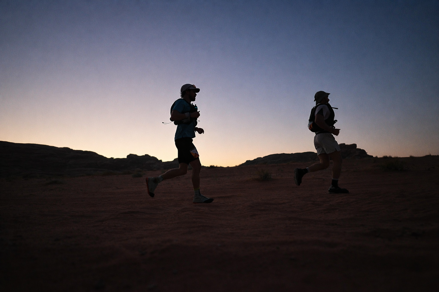 Twee mensen rennen door een woestijnachtig landschap bij schemering of zonsondergang, met silhouetten tegen de ondergaande zon.
