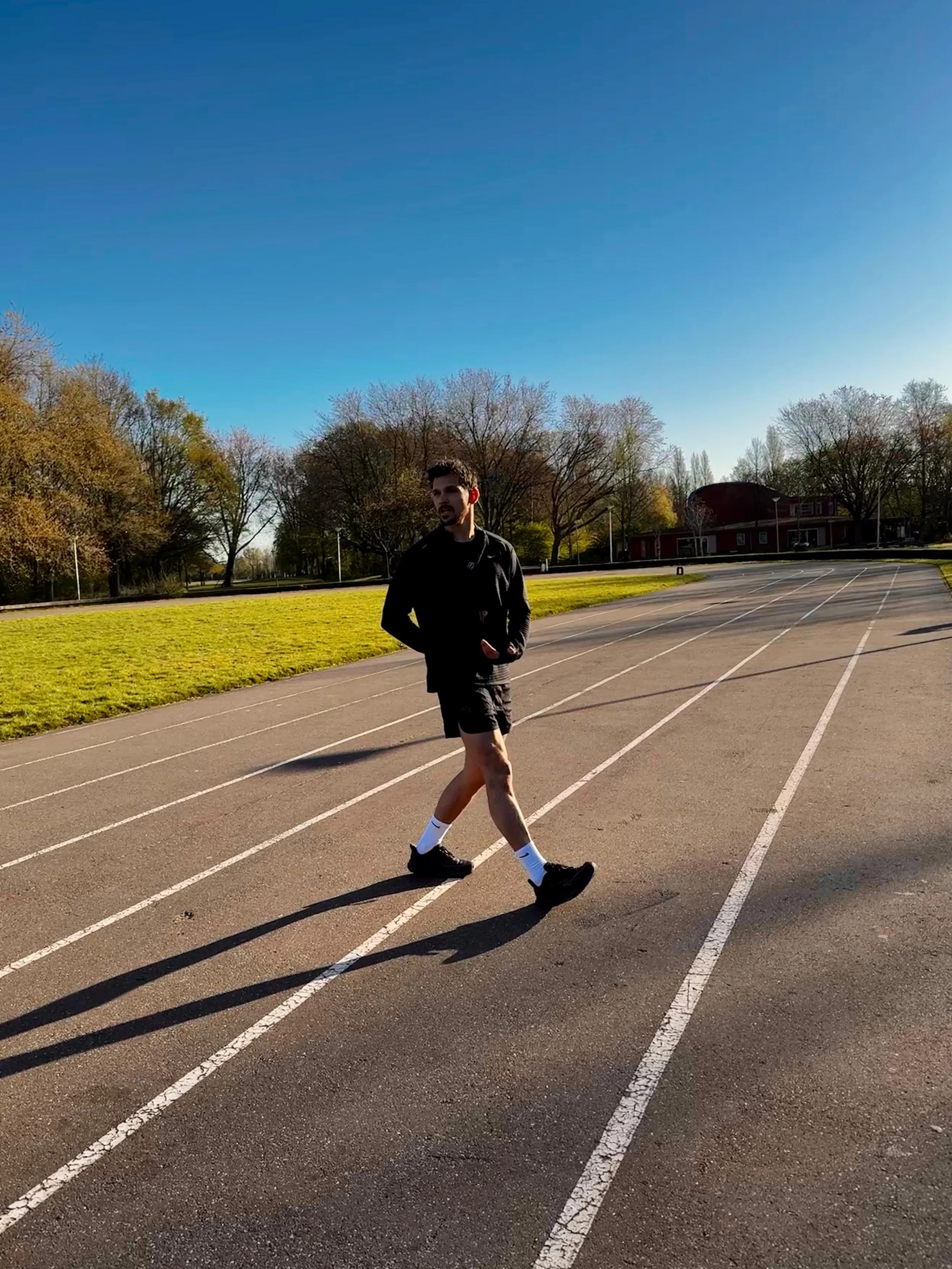Een man loopt op een atletiekbaan in de buitenlucht onder een heldere blauwe hemel, omringd door bomen.