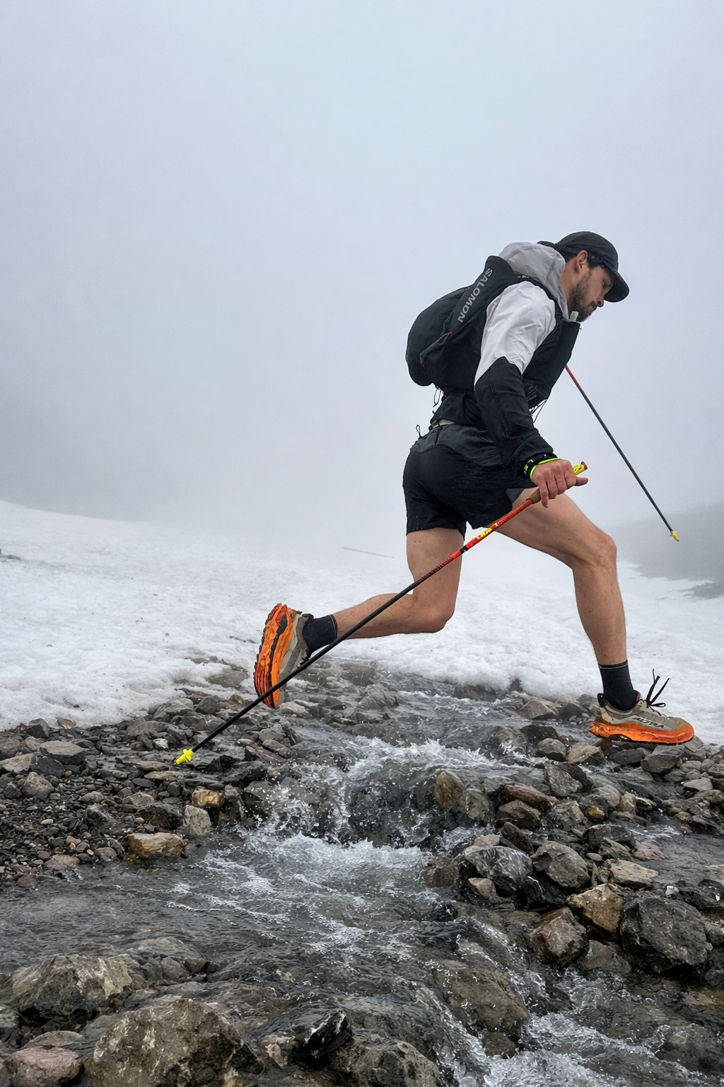 Een man in sportkleding en wandelschoenen loopt door een smal beekje in een bergachtig, mistig landschap, gebruikmakend van wandelstokken.