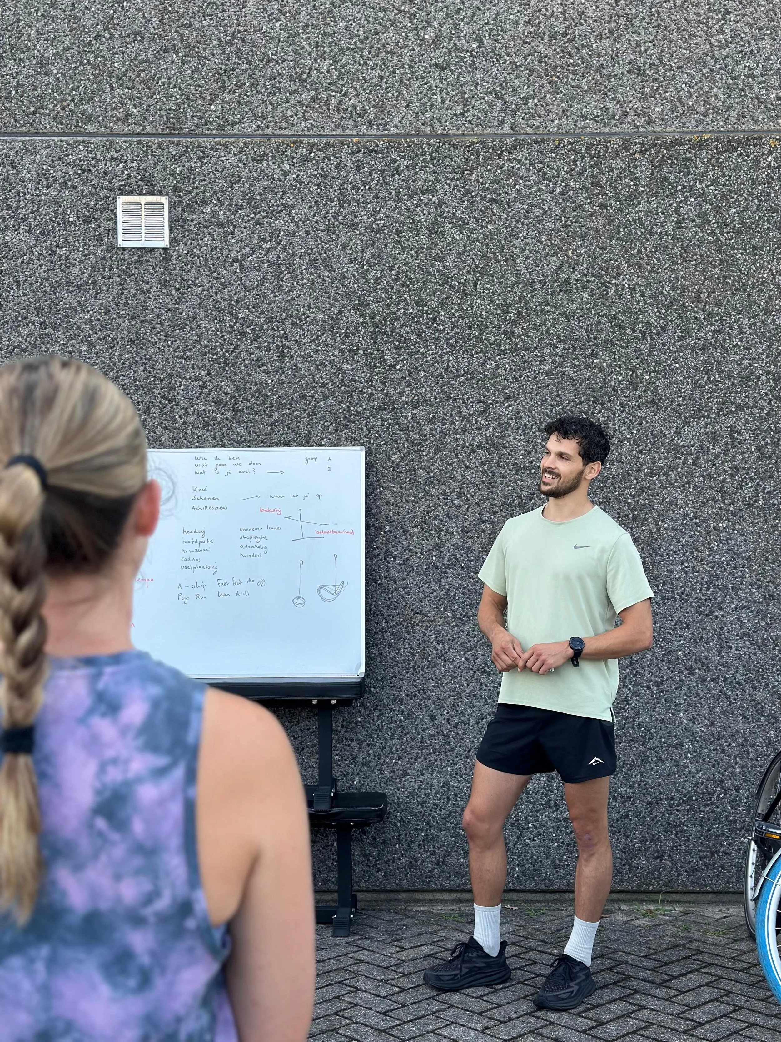Een man geeft een buitenles over fitness of sport aan een groep mensen, met een whiteboard vol aantekeningen en diagrammen.