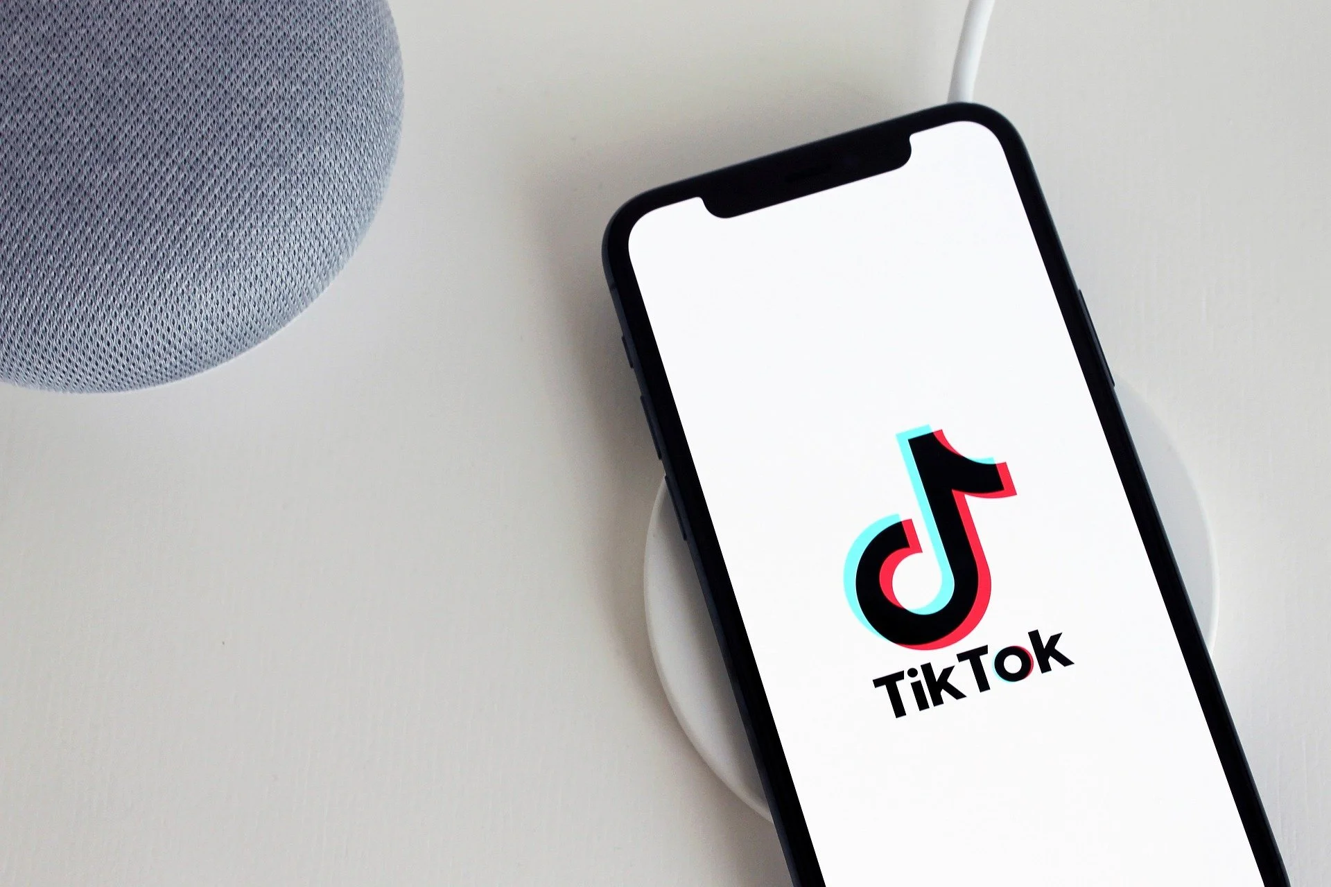 Smartphone displaying TikTok logo on a wireless charger next to a grey speaker on a white surface.