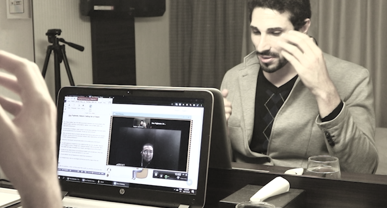 A man is sitting at a table in front of a laptop, engaging in a video call, with a third person visible on the laptop screen. The man has dark hair and a beard, and is wearing a light-colored blazer over a dark shirt. There is a glass of water and a white device on the table.