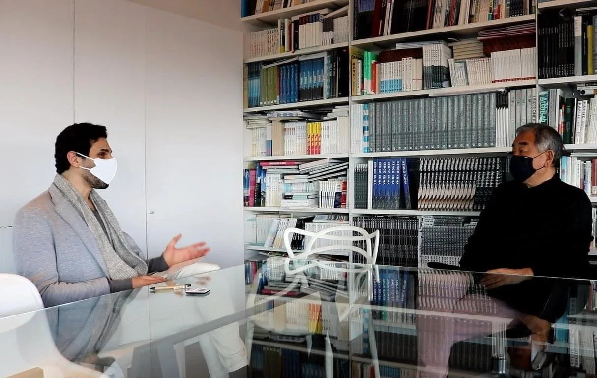 Two men wearing face masks sitting at a glass table in a room with a large bookshelf filled with books in the background, engaging in conversation.