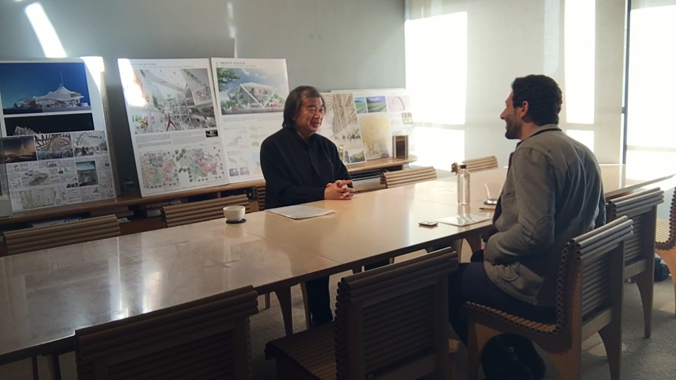 Two men sitting across from each other at a conference table in a room with display boards and architectural plans in the background, engaging in conversation.