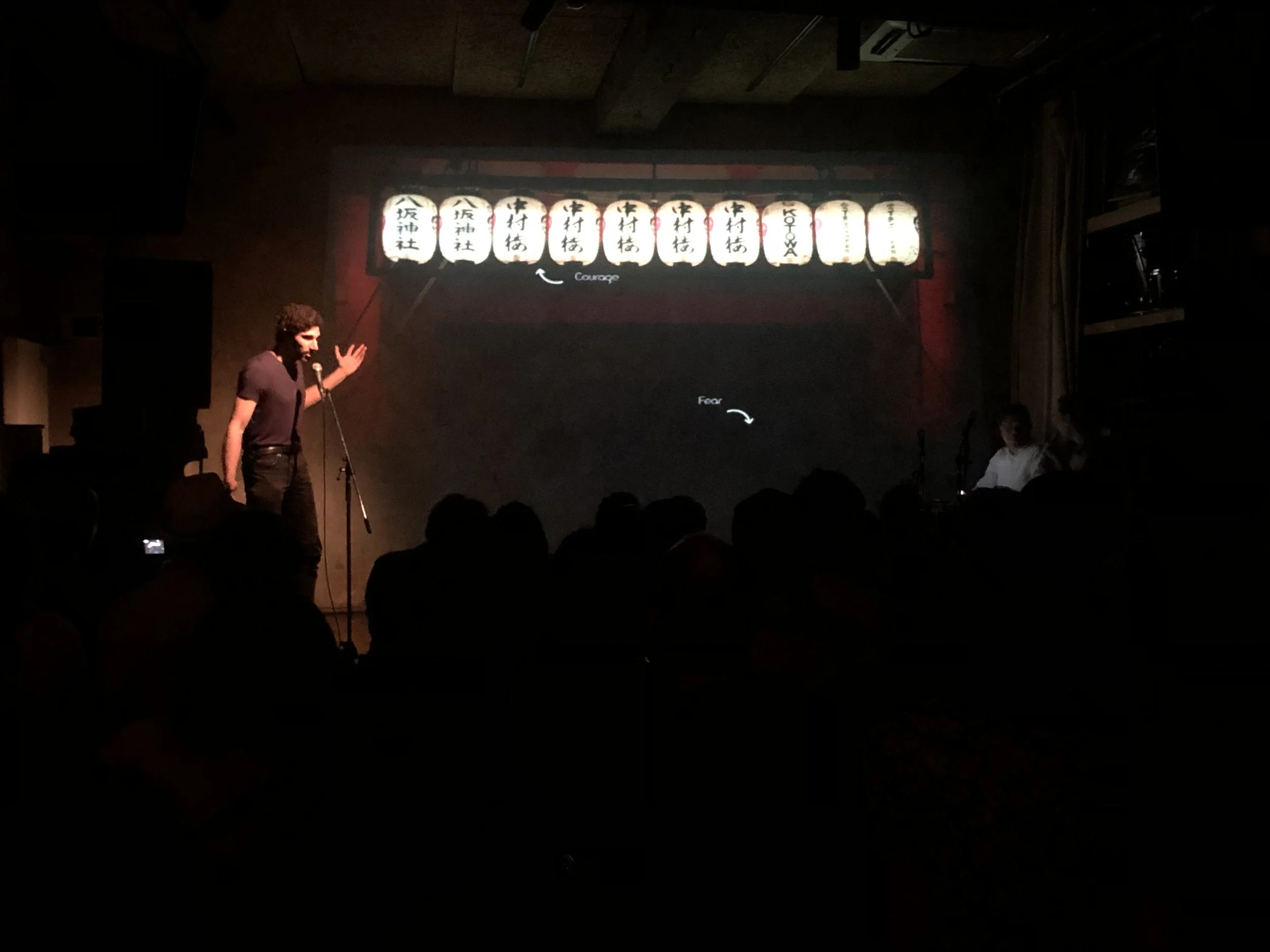 A person performing on stage with a microphone, facing an audience in a dimly lit room. A projected slide with Japanese lanterns and annotations about courage and fear is visible behind them.
