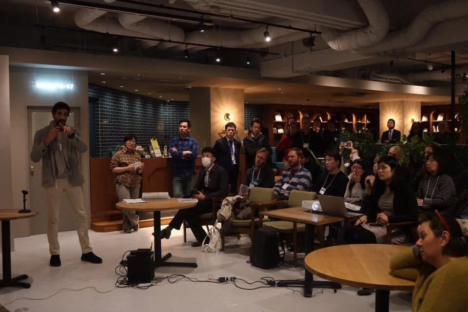A man standing on the left is speaking into a microphone to an audience seated in a modern, dimly lit room, with a projector on a table in front of him, and several attendees taking notes or recording.