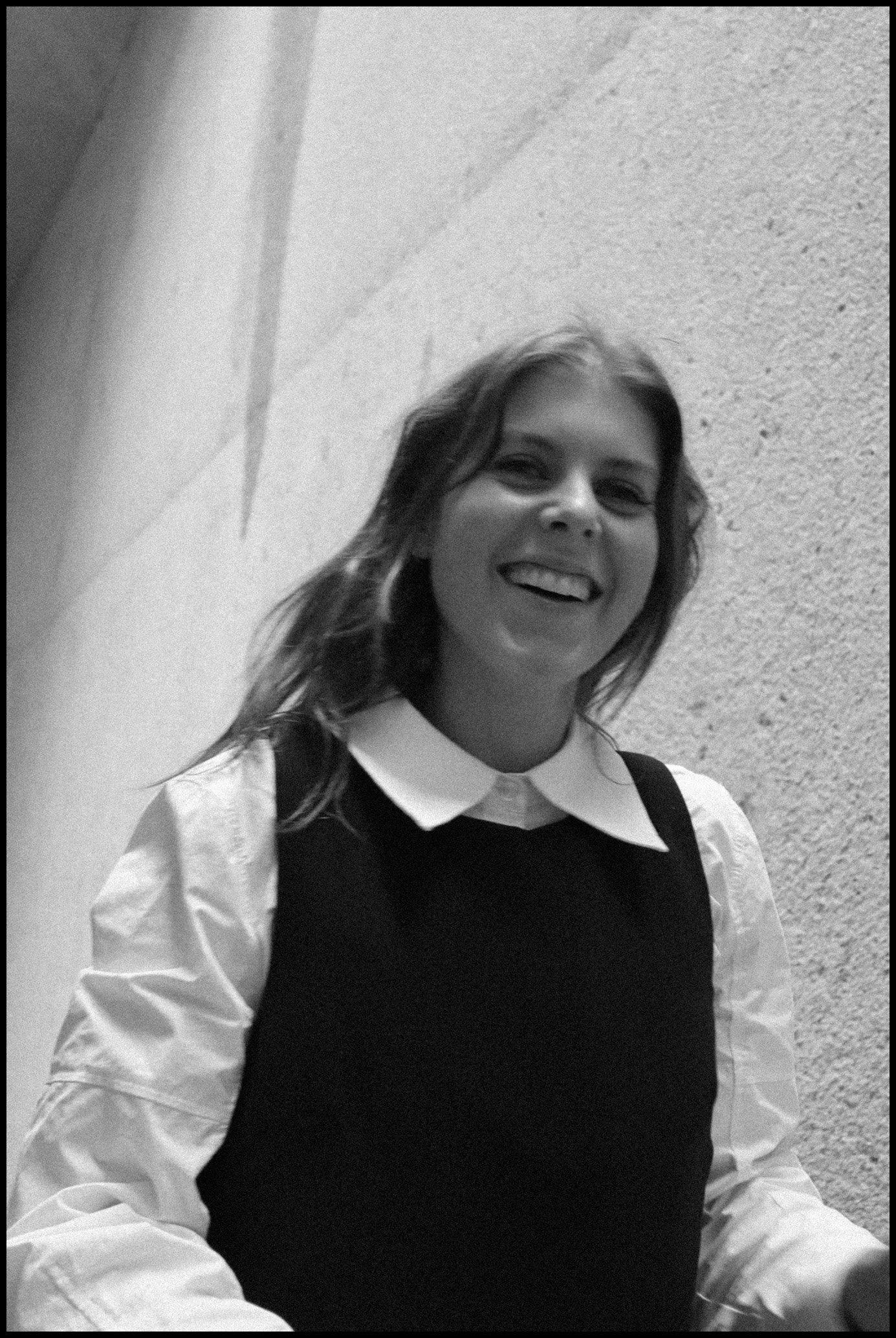 A woman with shoulder-length hair smiling, dressed in a collared shirt under a sleeveless top, standing against a textured wall.