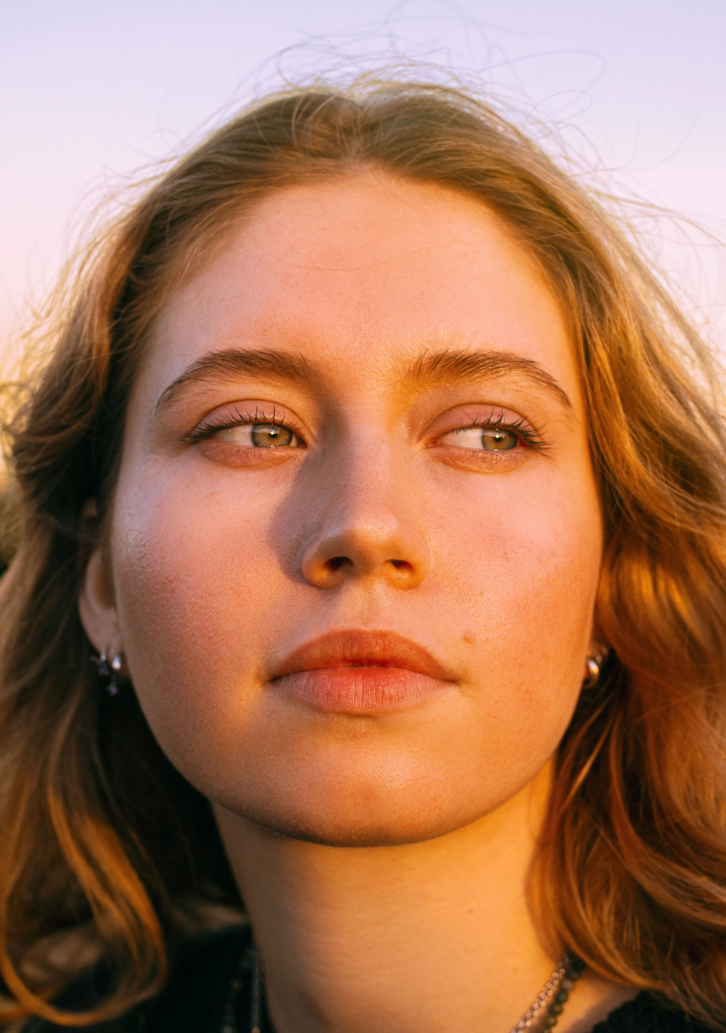 A close-up of a young woman with wavy hair outdoors during golden hour, looking off to the side with a serene expression.