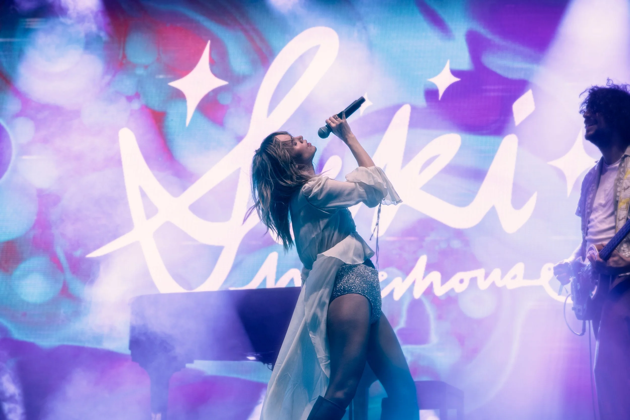 A woman singing on stage with a microphone, wearing sparkly shorts and a light-colored dress shirt. A man with curly hair and glasses plays guitar beside her. The background displays colorful lights and a large, stylized word or signature.