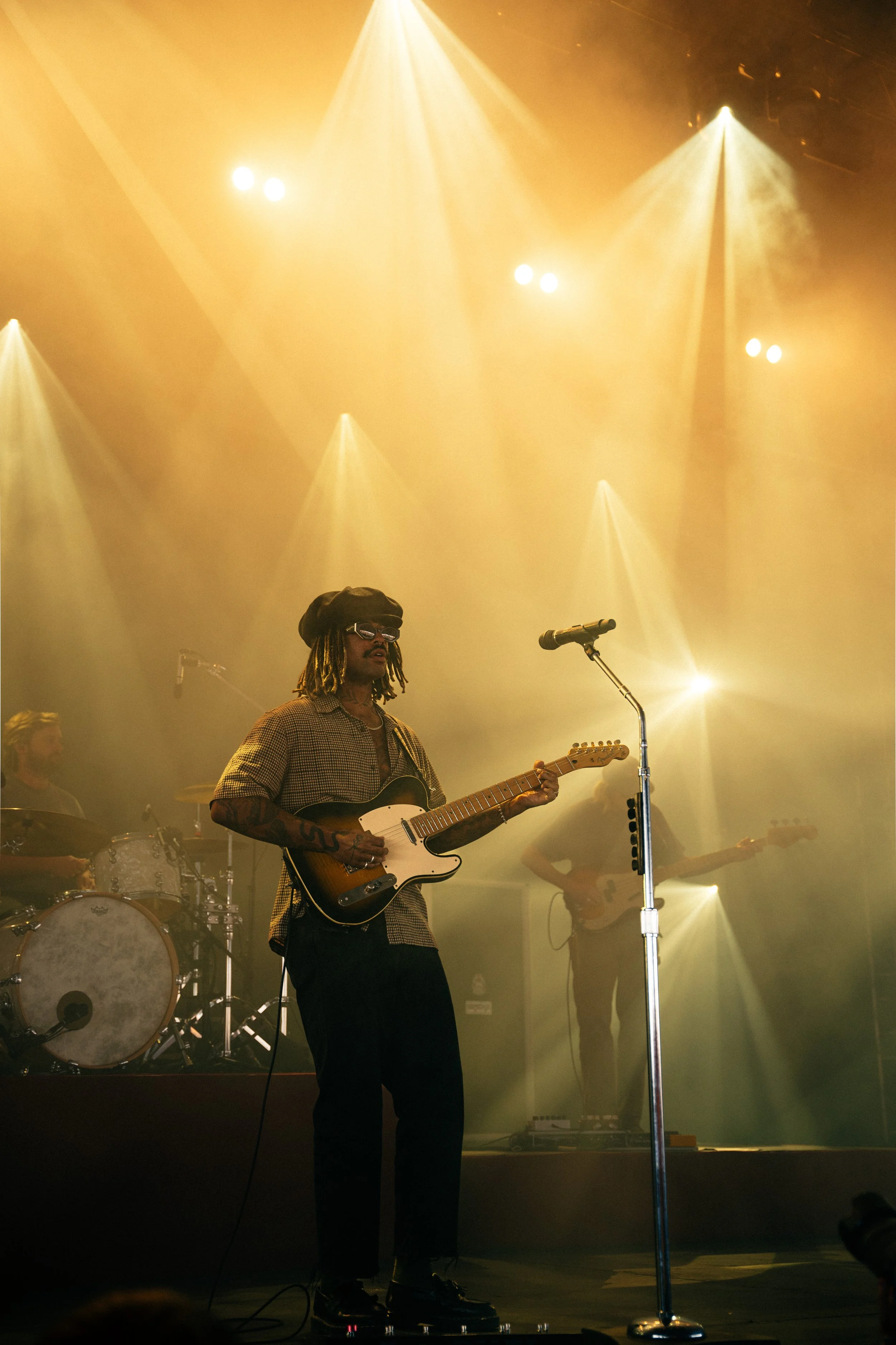 Musician playing an electric guitar on stage with dramatic lighting, backup drummer in the background, and another guitarist to the right.