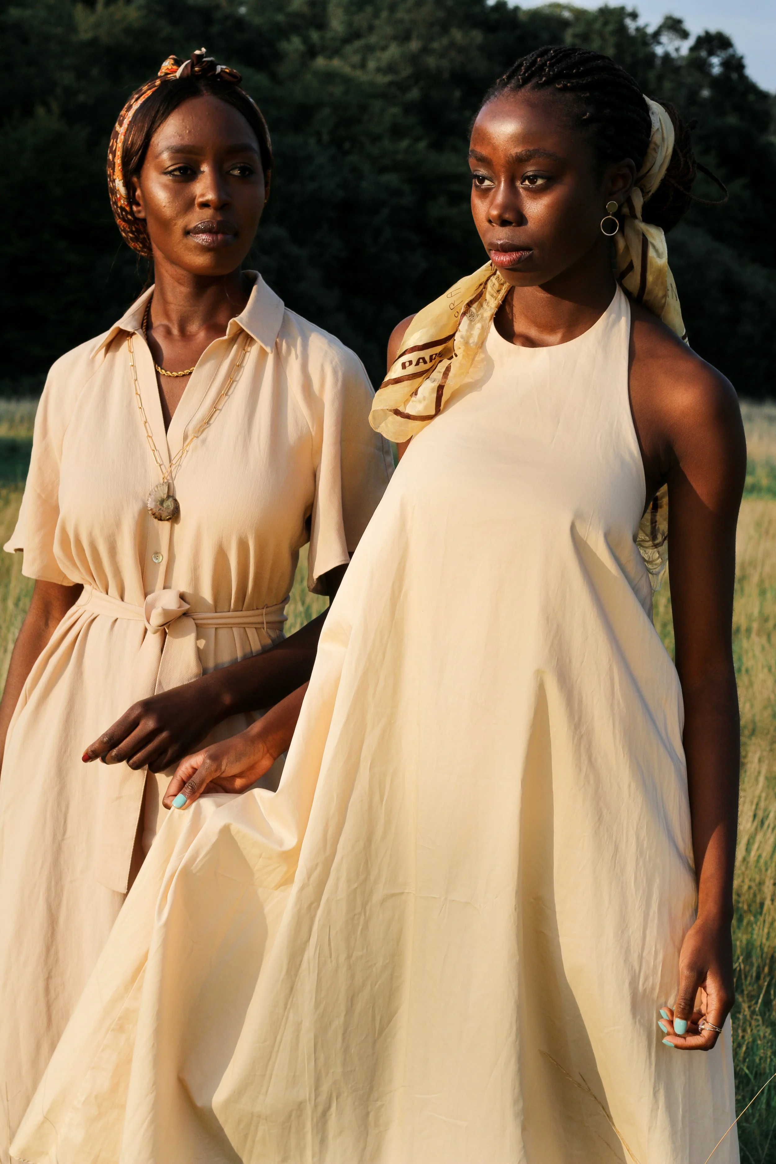Two women standing outdoors in a grassy field, wearing cream-colored dresses, with one woman holding the other's hand.