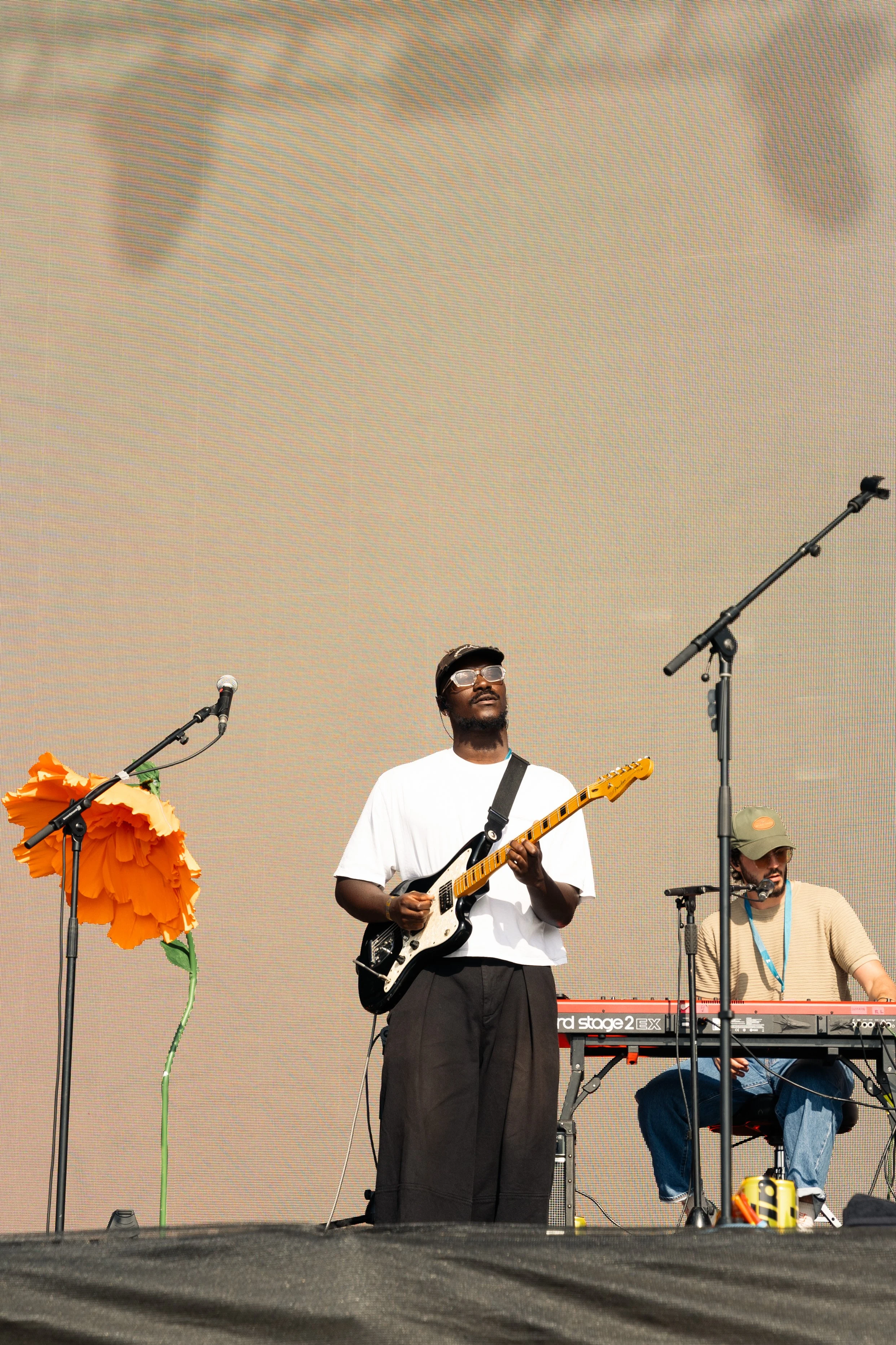 Two musicians performing on stage, one playing an electric guitar and the other on a keyboard. There is a large orange flower prop to the left of the guitarist.