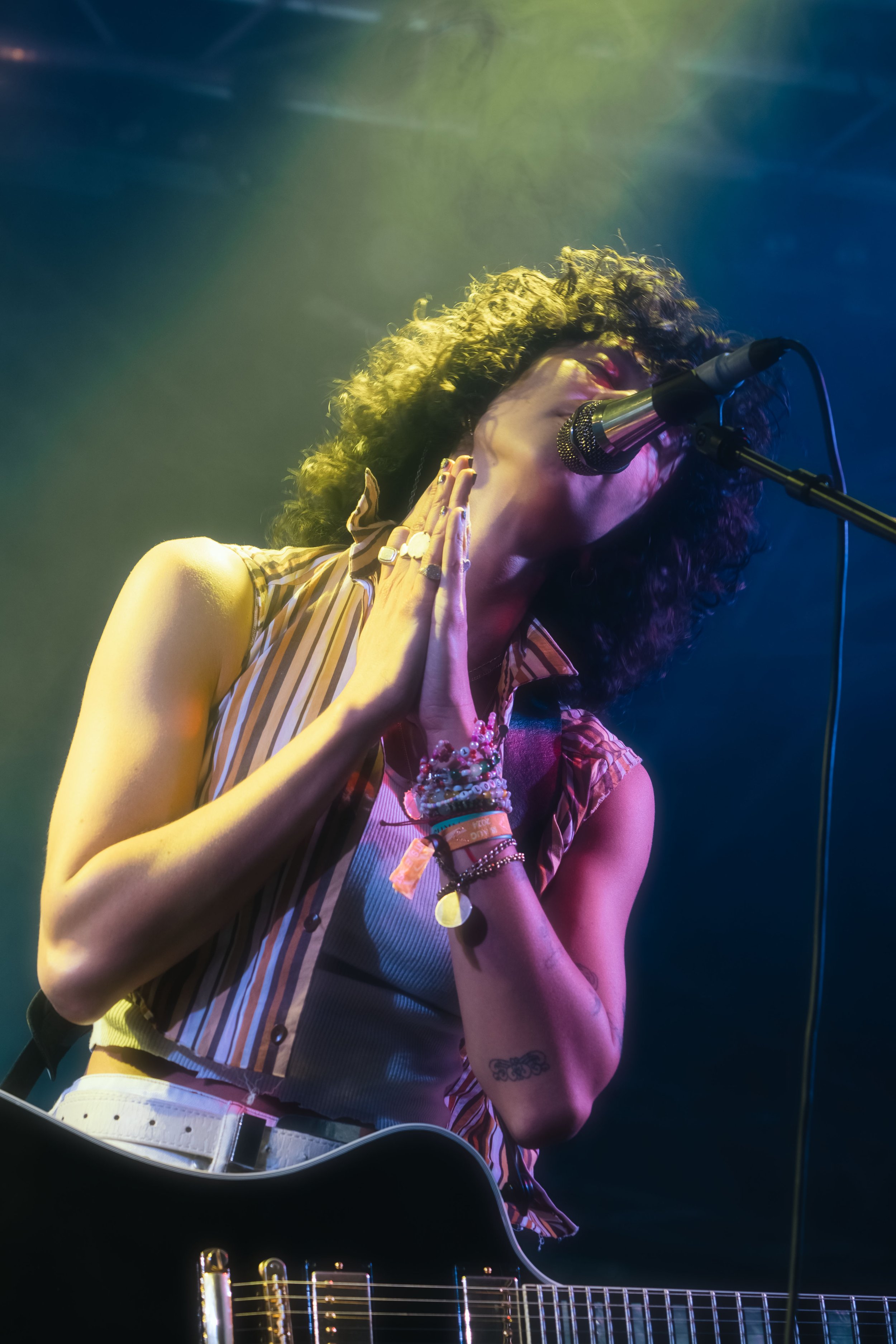 A woman singing into a microphone while playing an acoustic guitar on stage, illuminated by colored stage lights.