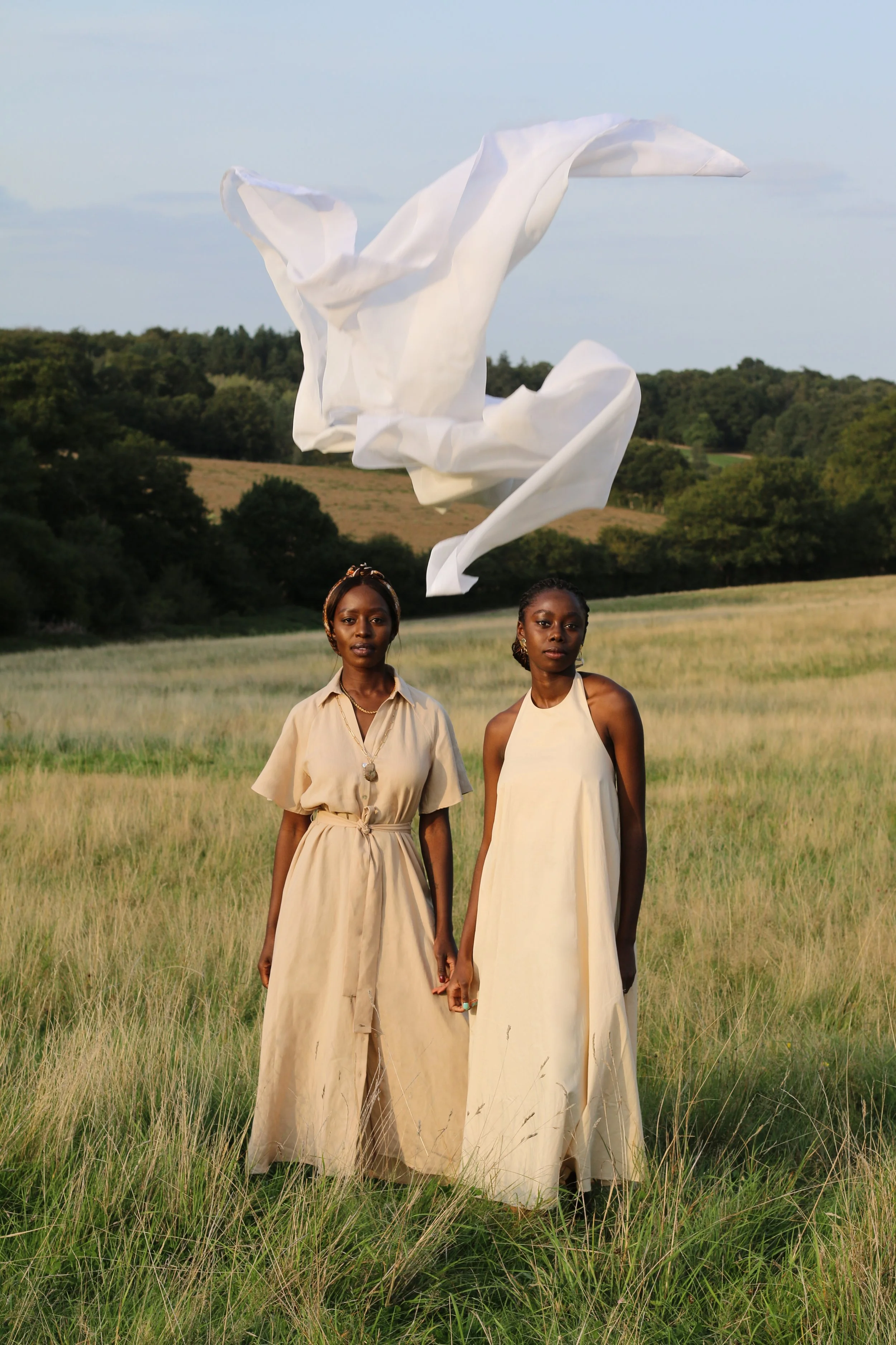 Two women standing in a grassy field, holding hands, with a piece of white fabric flying above them.