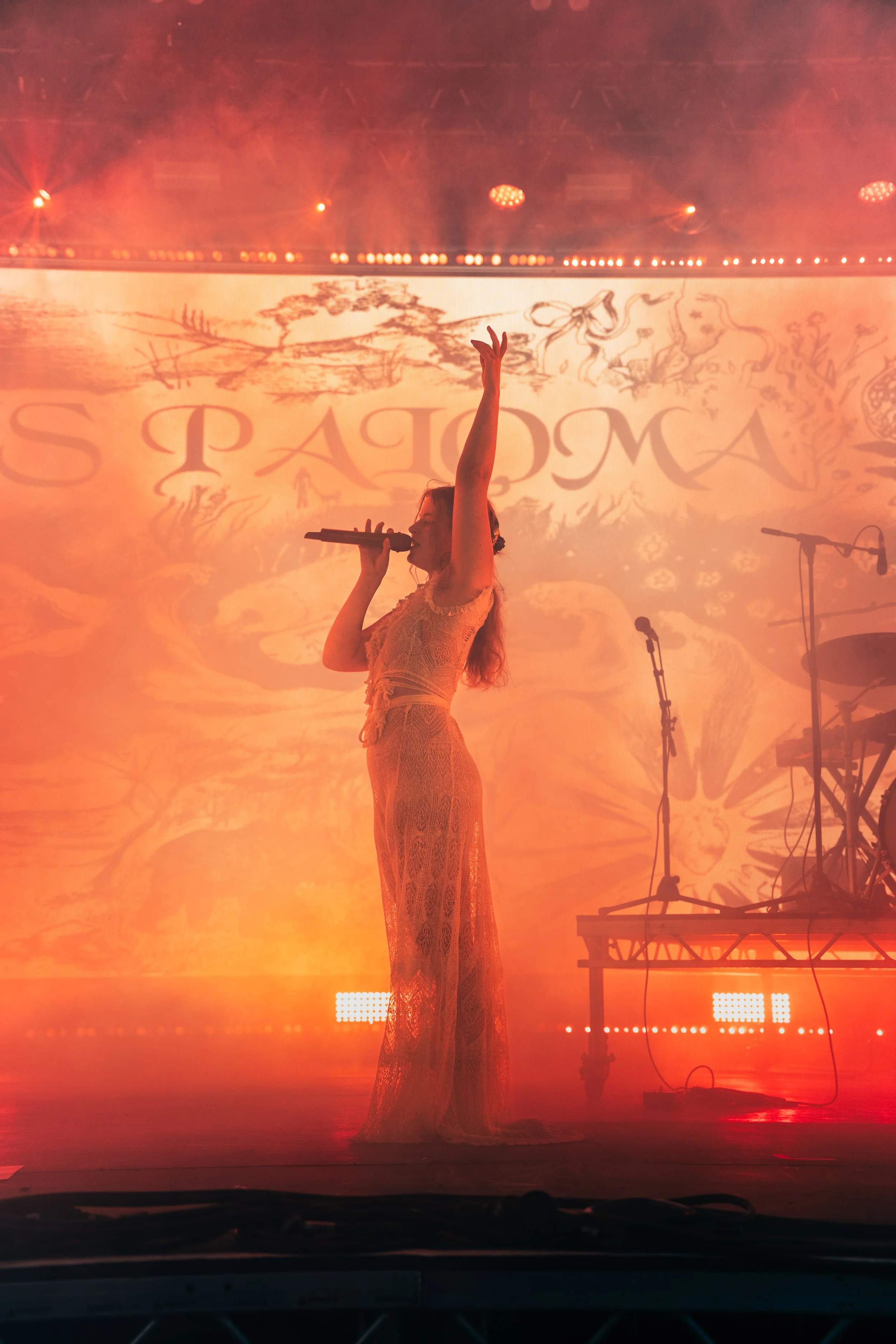 A female singer performing on stage with orange lighting, holding a microphone and raising one arm, with a backdrop displaying artistic illustrations and the word 'SPAIOMA'.