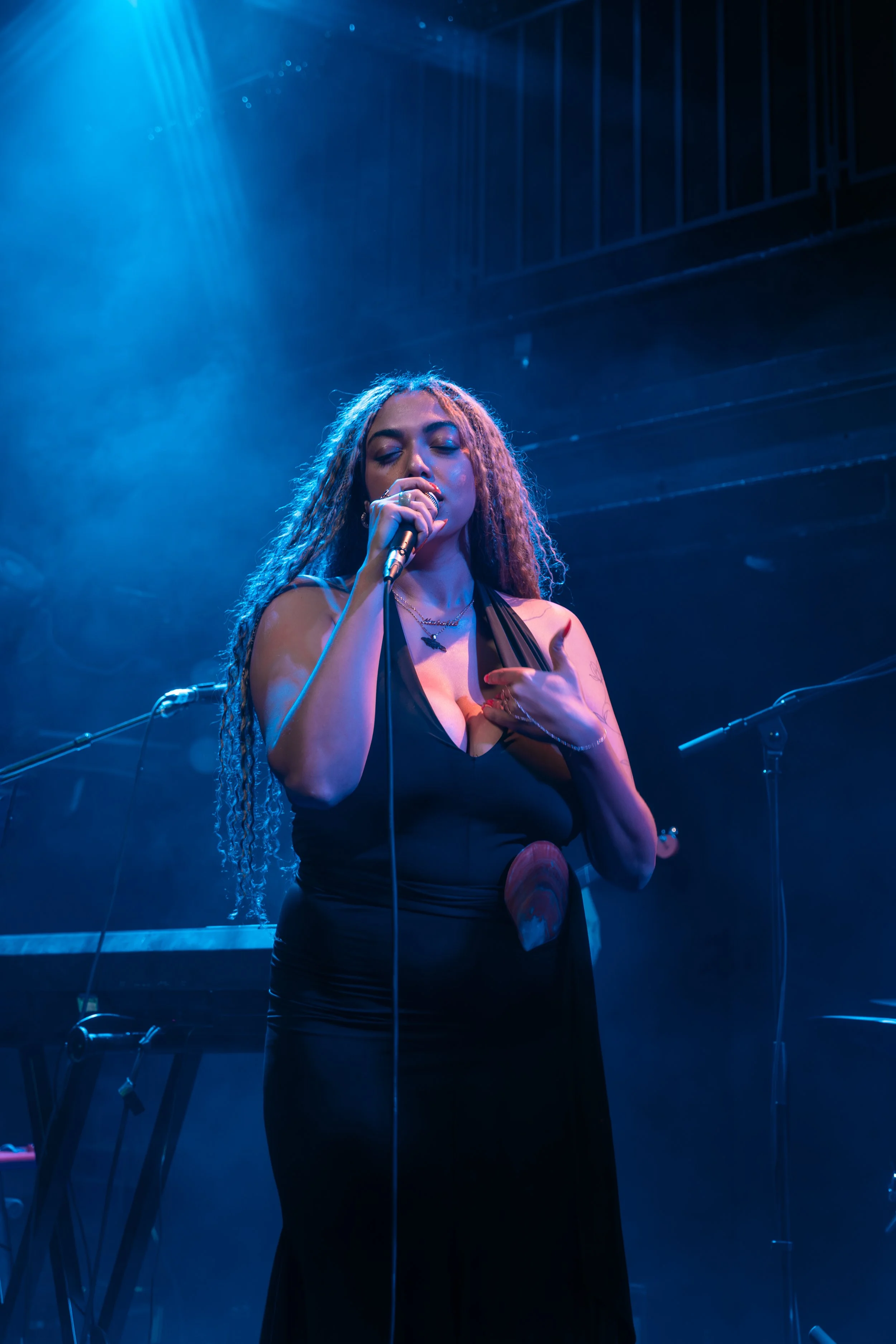 A female singer with long curly hair performing on stage, holding a microphone with her eyes closed, wearing a black dress, illuminated by stage blue lighting.